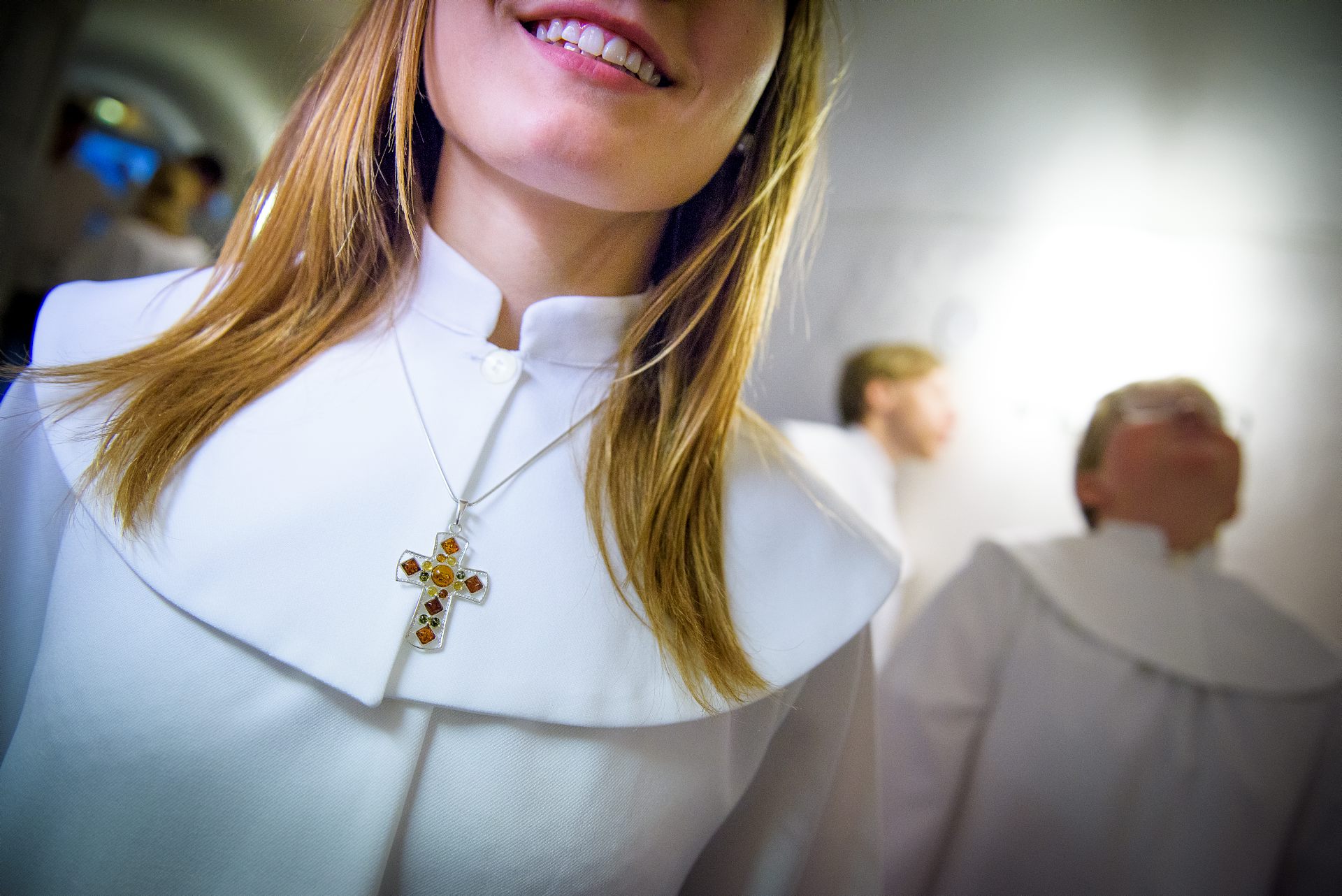 En närbild på ett halssmycke i form av ett kors runt halsen på en leende konfirmand i kåpa