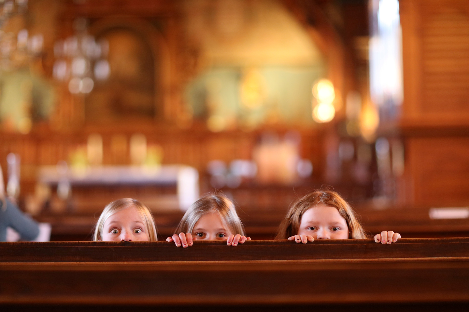 Tre flickor tittar upp bakom en kyrkbänk.