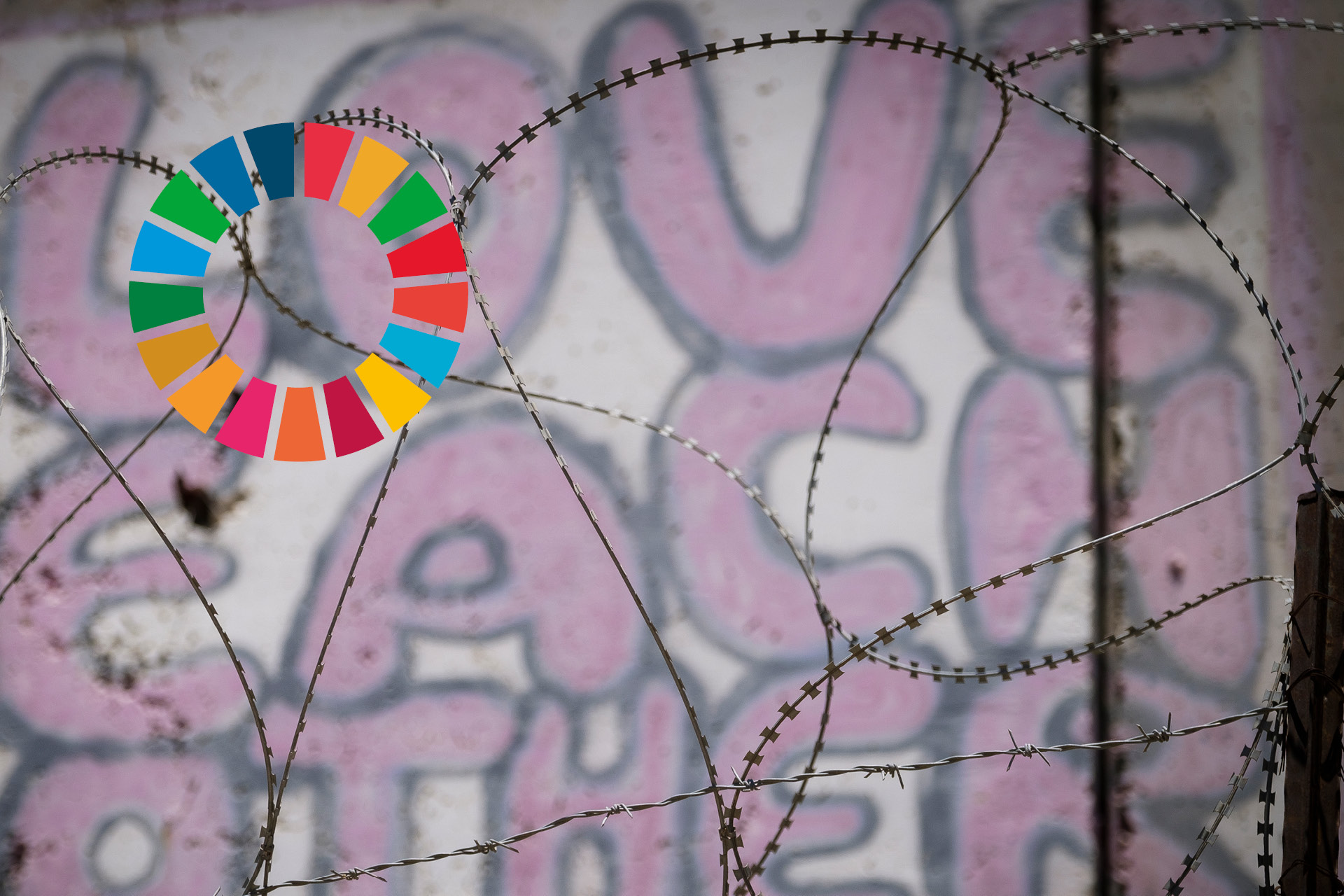 Grafitti på en vägg, med texten Love each other. I förgrunden taggtråd. Till vänster i bilden syns loggan för Agenda 2030.