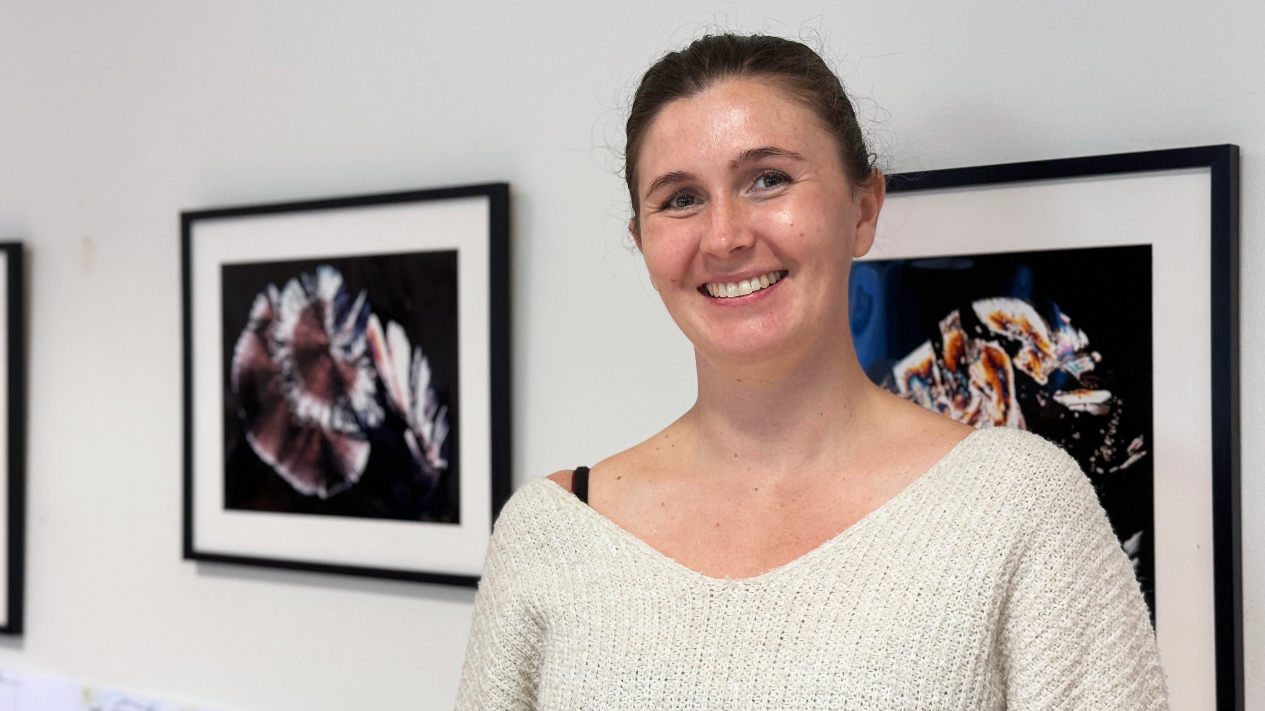 En ung kvinna ler framför en vägg med tavlor.