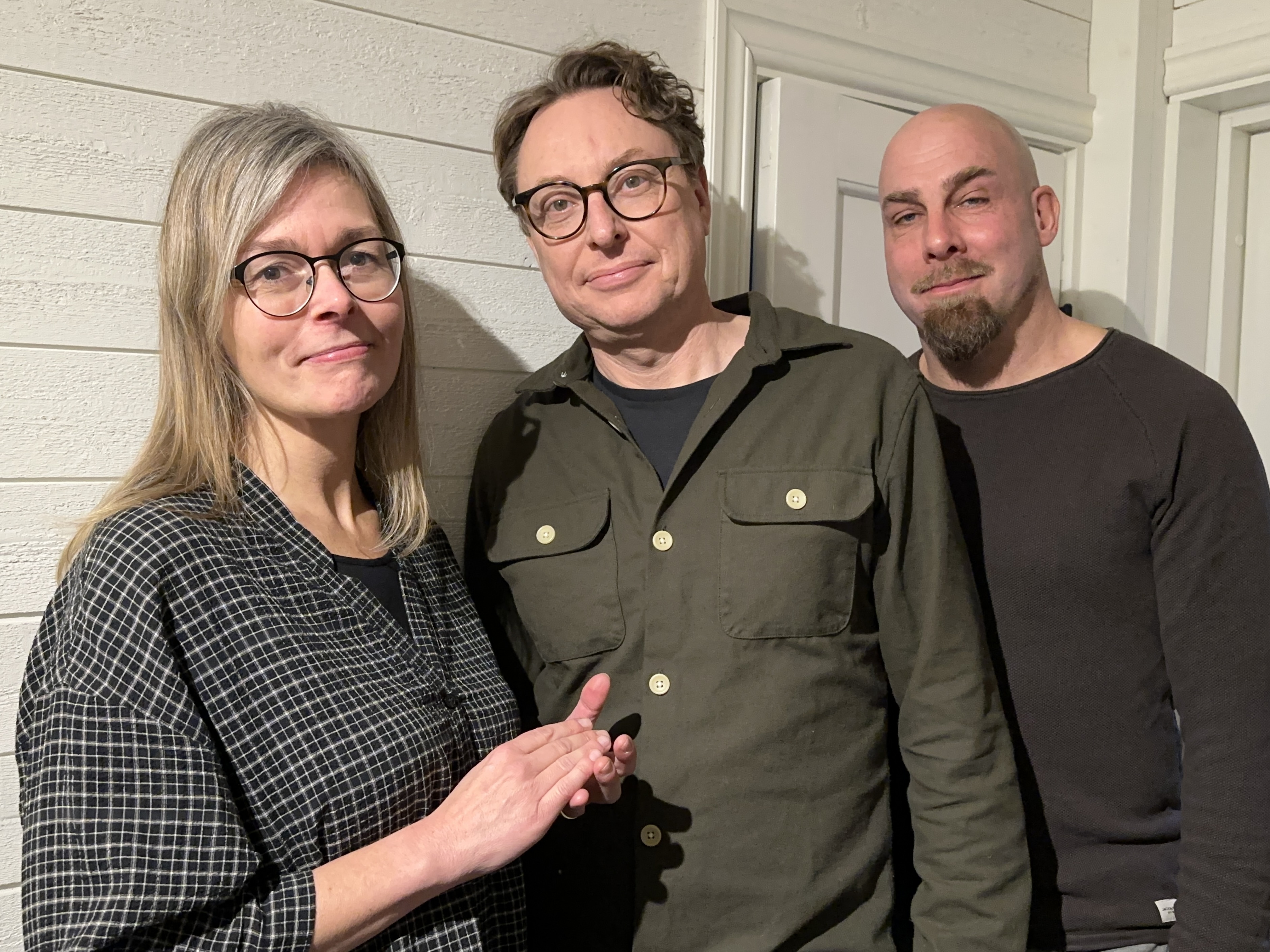 Tre personer, en kvinna och två män står tillsammans framför en vit vägg