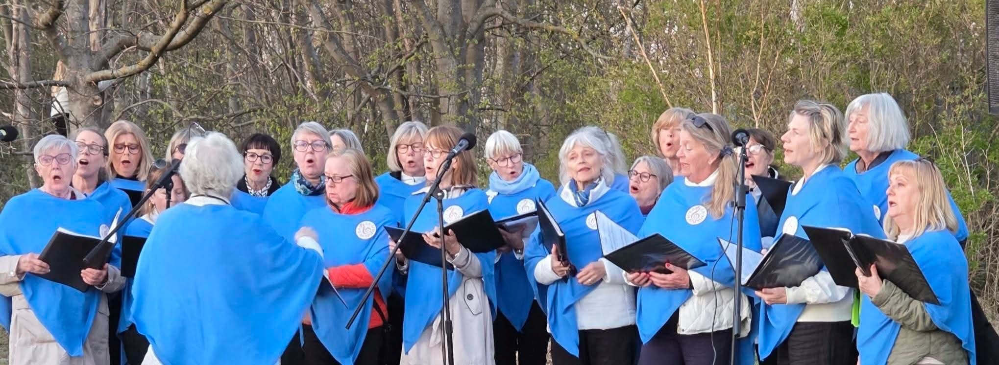 Grupp av kvinnor stående utomhus. De har blå halvkåpor på sig och håller svarta pärmar i händerna.