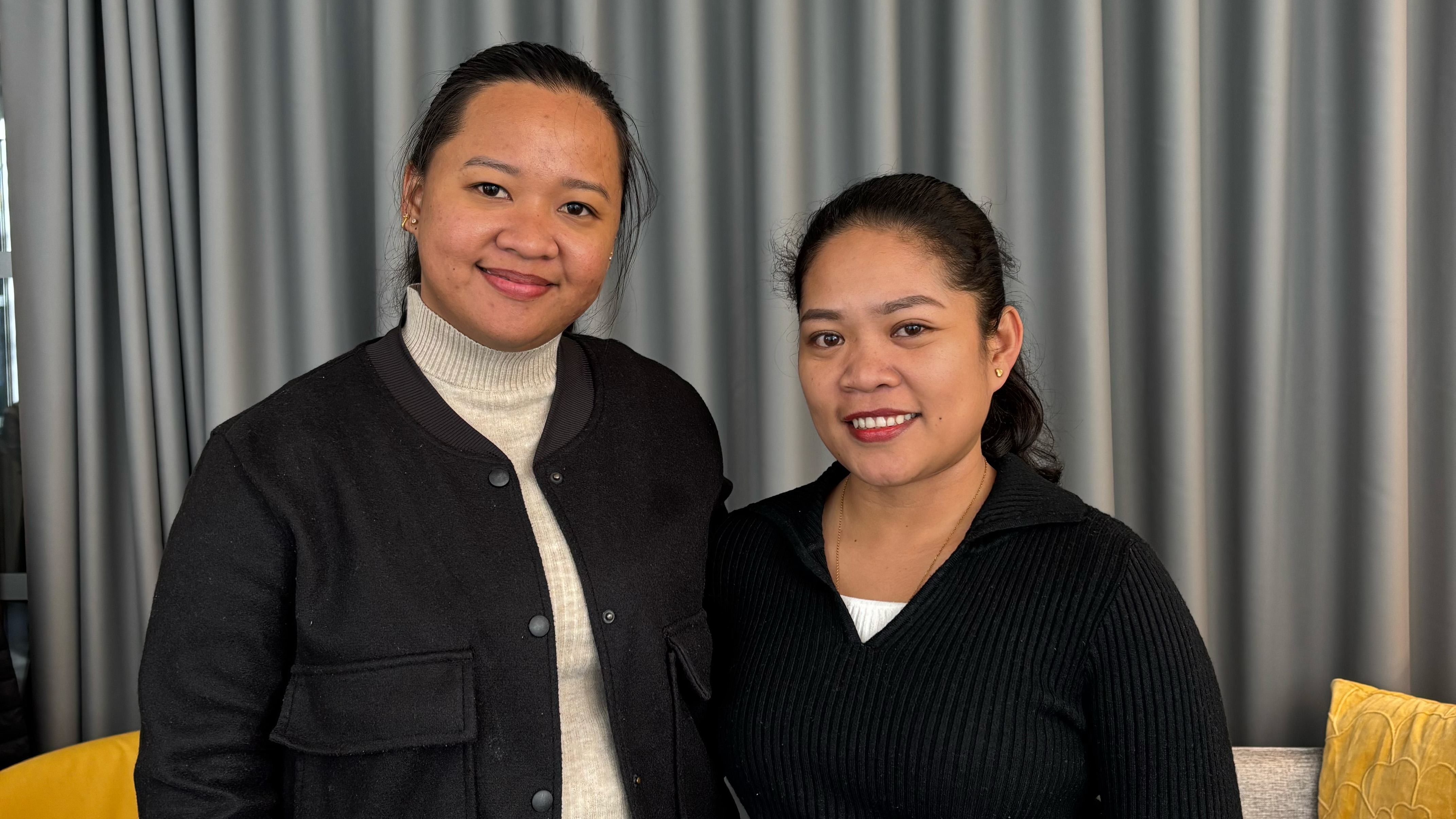 Två kvinnor stående framför en grå vägg