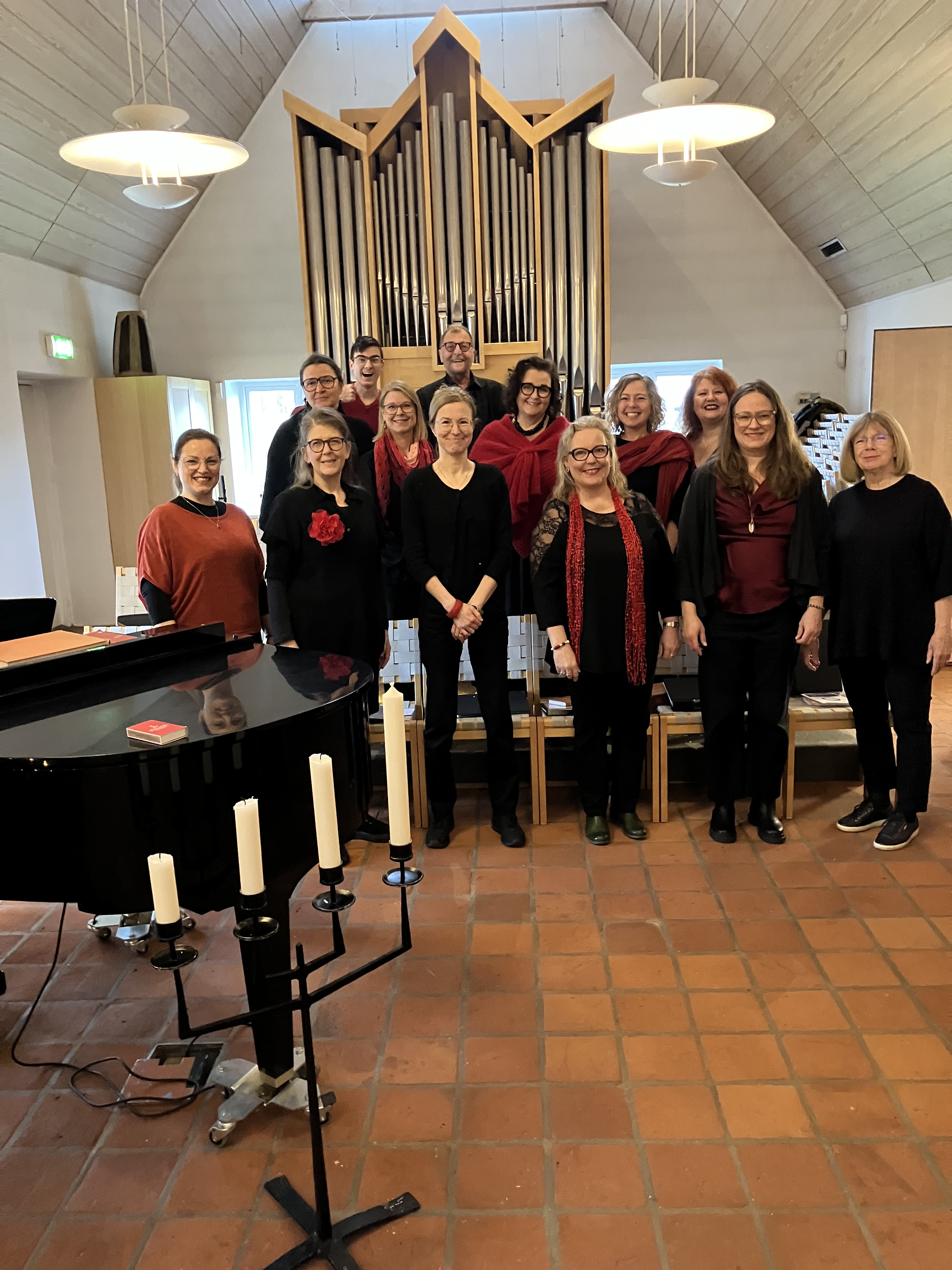 Mindre grupp av kvinnor och män klädda i rött och svart stående bakom en svart flygel men framför en orgel. Alla ser glada ut.