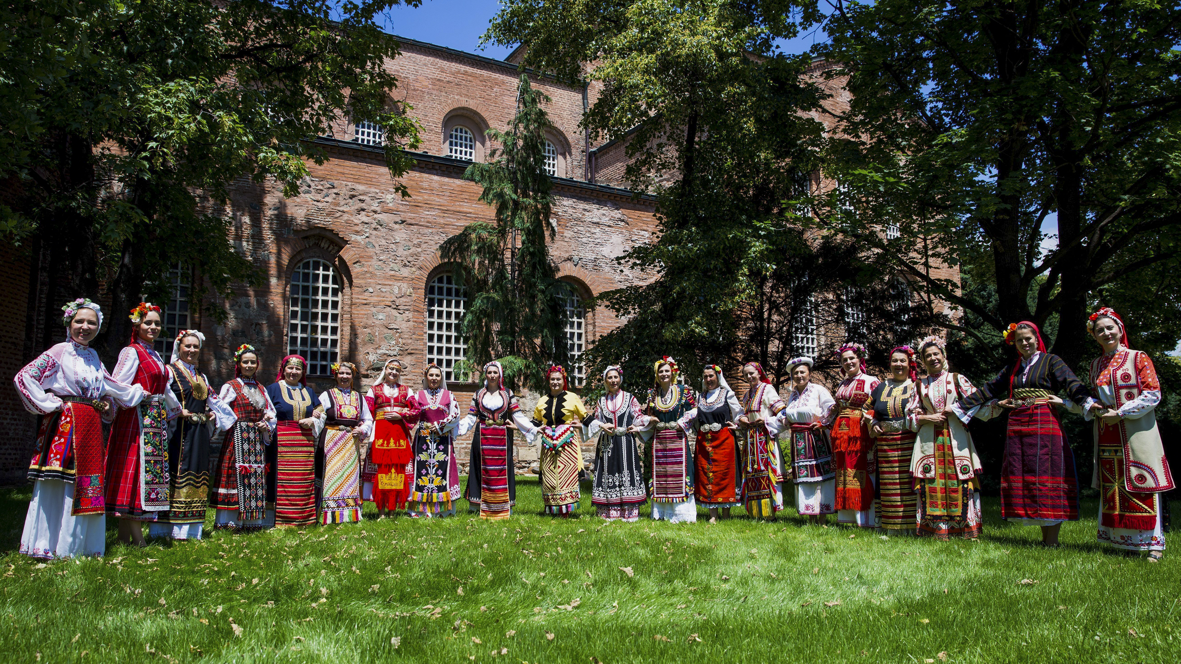 Tjugo kvinnor i traditionella balkiska folkdräkter står uppställda i halvmåne på en gräsmatta framför en röd tegelbyggnad.