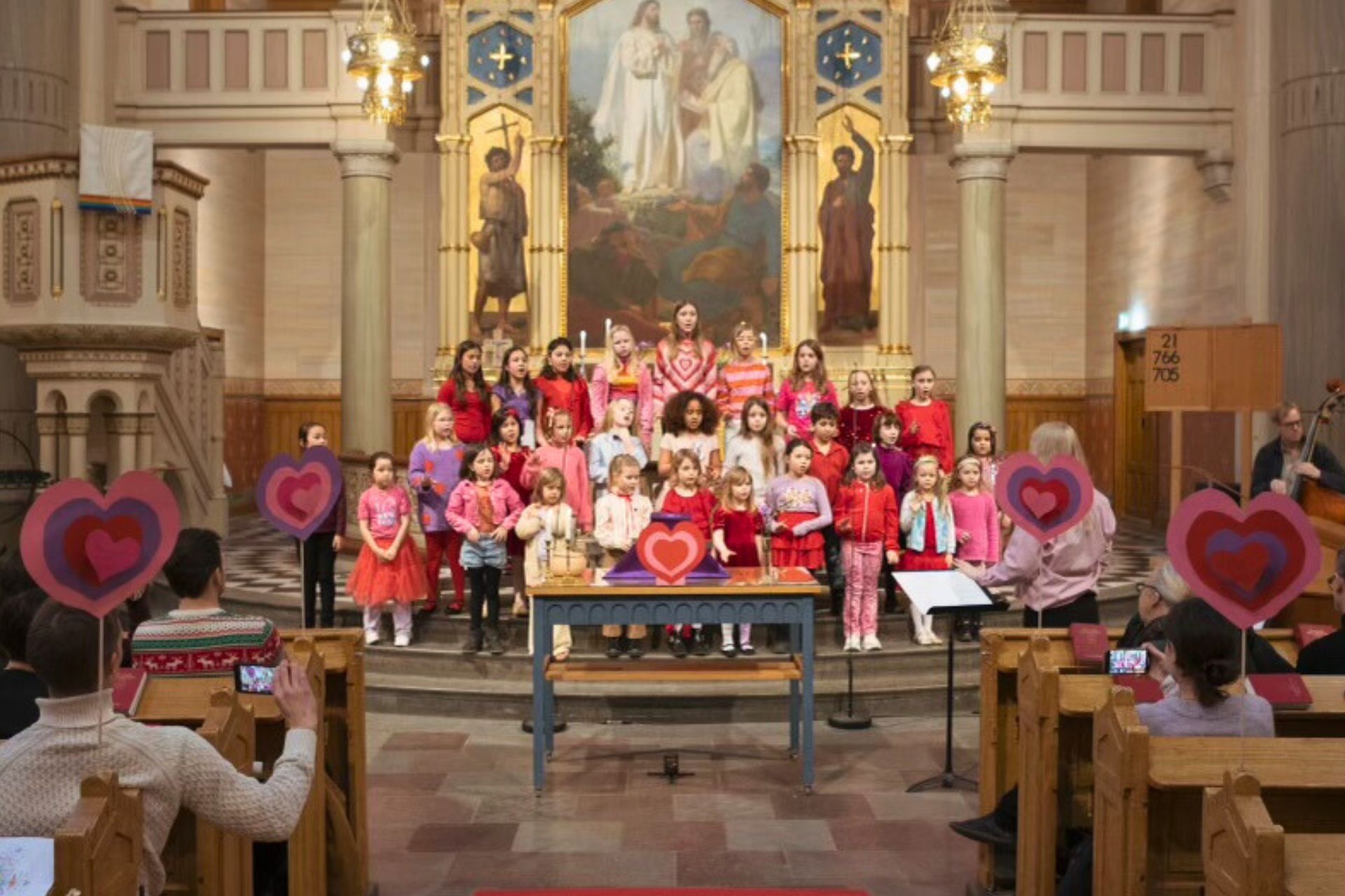 Bild av en barnkör som sjunger i kyrkan. Alla är rödklädda och det finns dekorationer med hjärtan runt omkring.