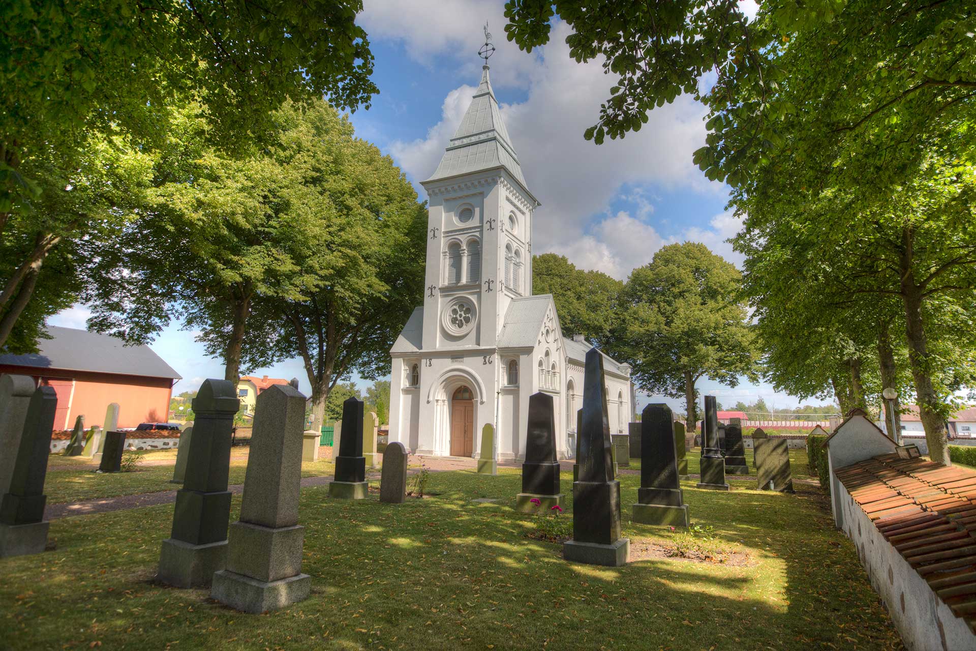 Vit 1800-talskyrka i strålande sol, omgiven av lummiga träd.