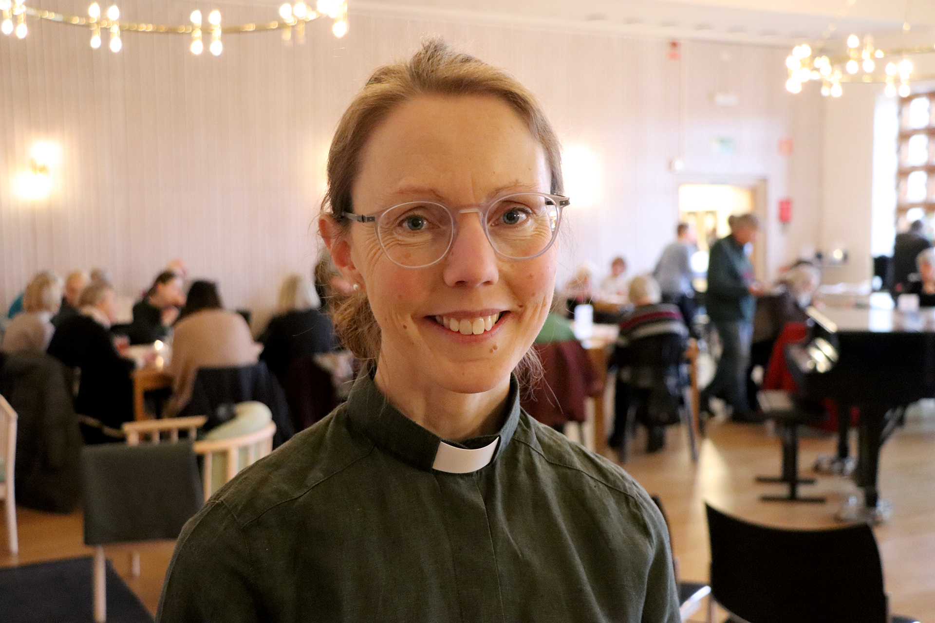 Ett foto på Maria i grön diakonskjorta, i bakgrunden syns människor som sitter vid bord. 