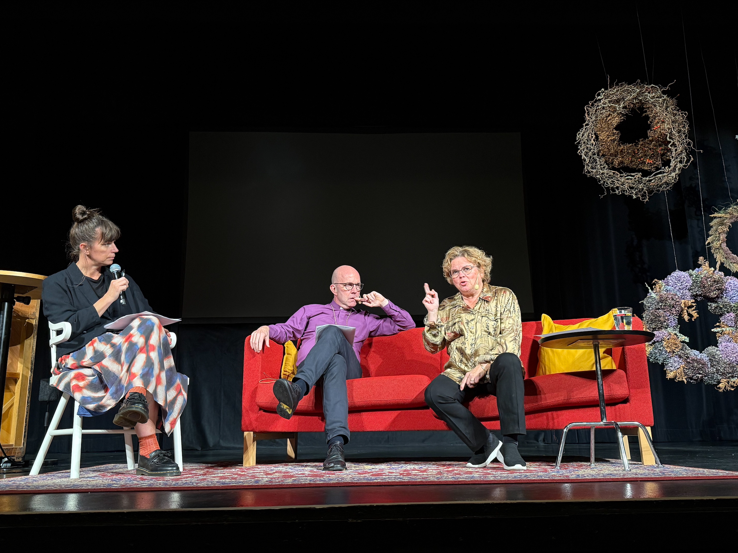 Tre personer sitter på en scen, Isabelle McCallister sitter längst till vänster på en stol. I en röd soffa sitter biskop Fredrik Modéus och Maria Wolrath Söderberg.