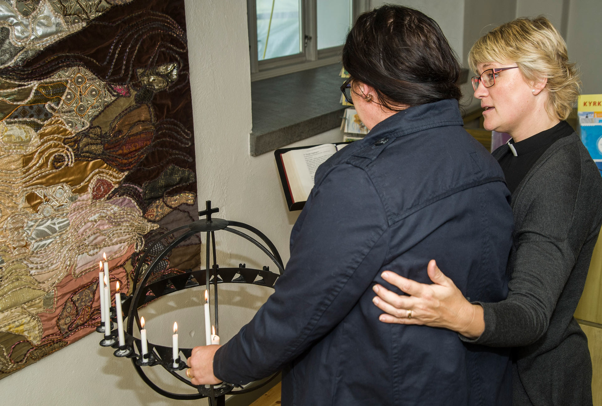 En präst tröstar en person som precis tänt ett ljus för att sätta i ljusbäraren i en kyrka.