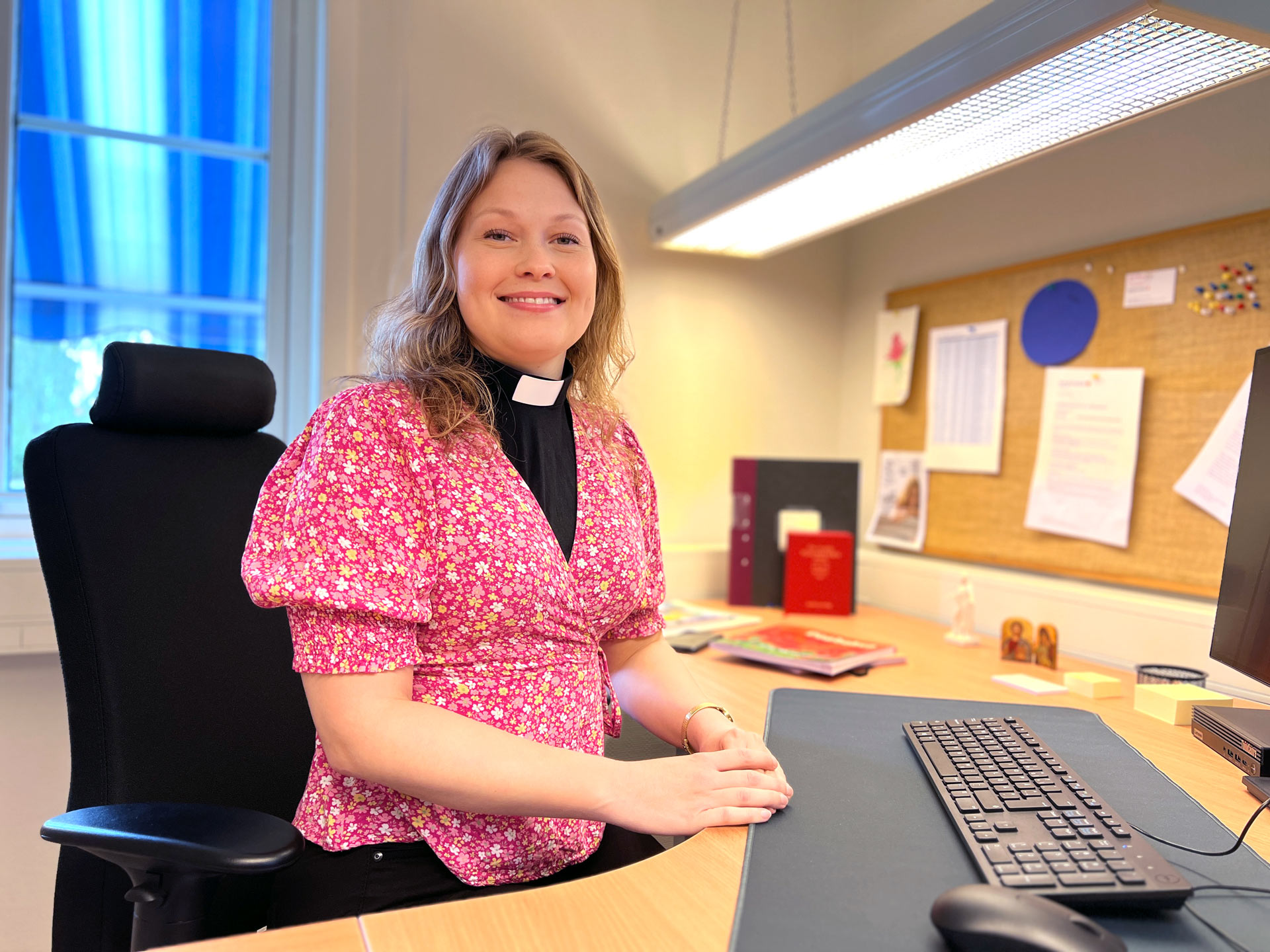 En ung kvinna sitter vid vid ett skrivbord framför ett tangentborden och dataskärm. Kvinnan har en blommig rosa blus över en svart prästskjorta med prästkrage.