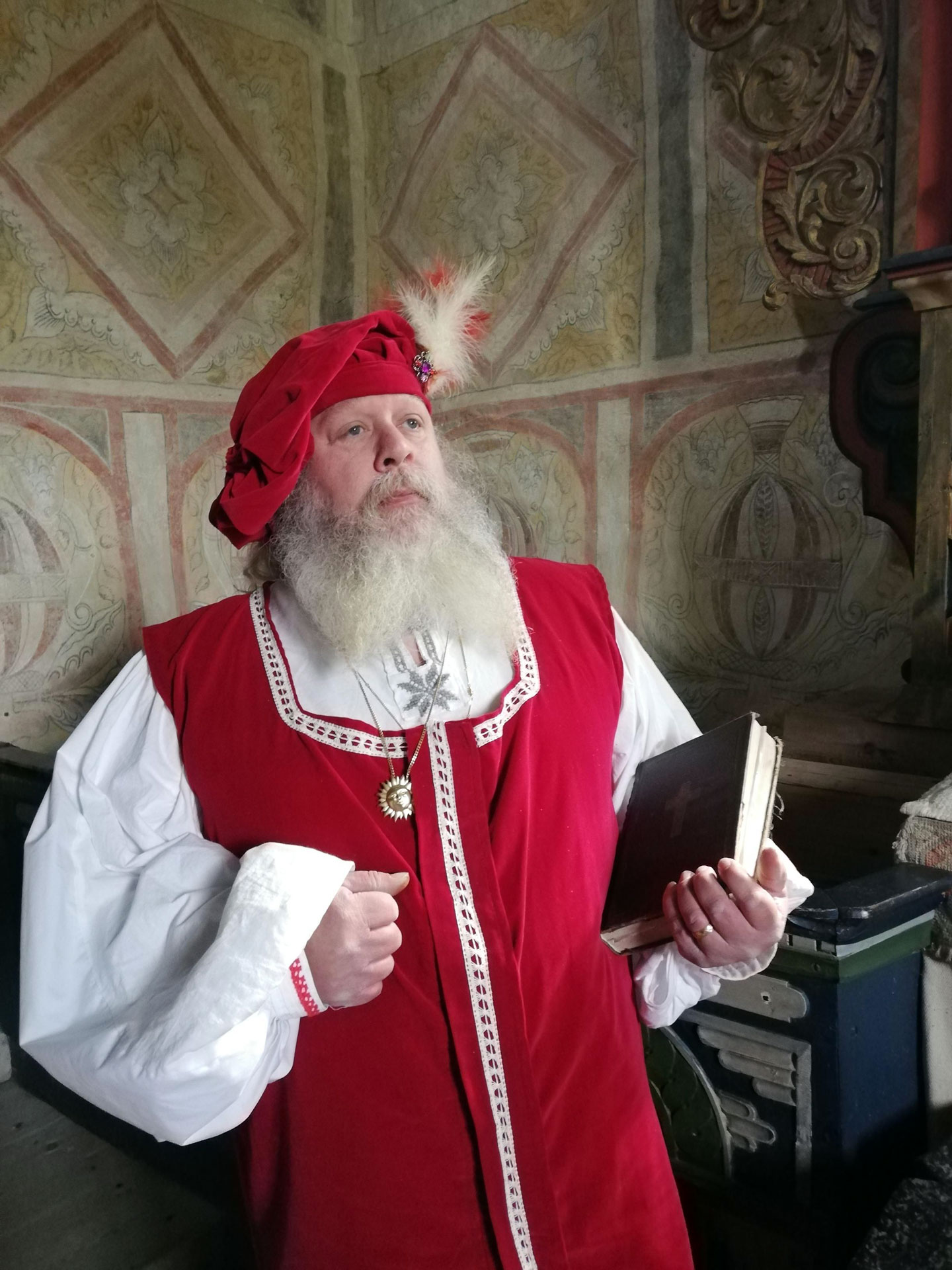 En man med stort vitt skägg. Mannen bär medeltidskläder. en skjorta med vida puffärmar, röd väst och en baskerliknande huvudbonad med fjäder. I handen håller han en bibel. I bakgrunden ser vi väggmålningar i från en medeltida kyrka.