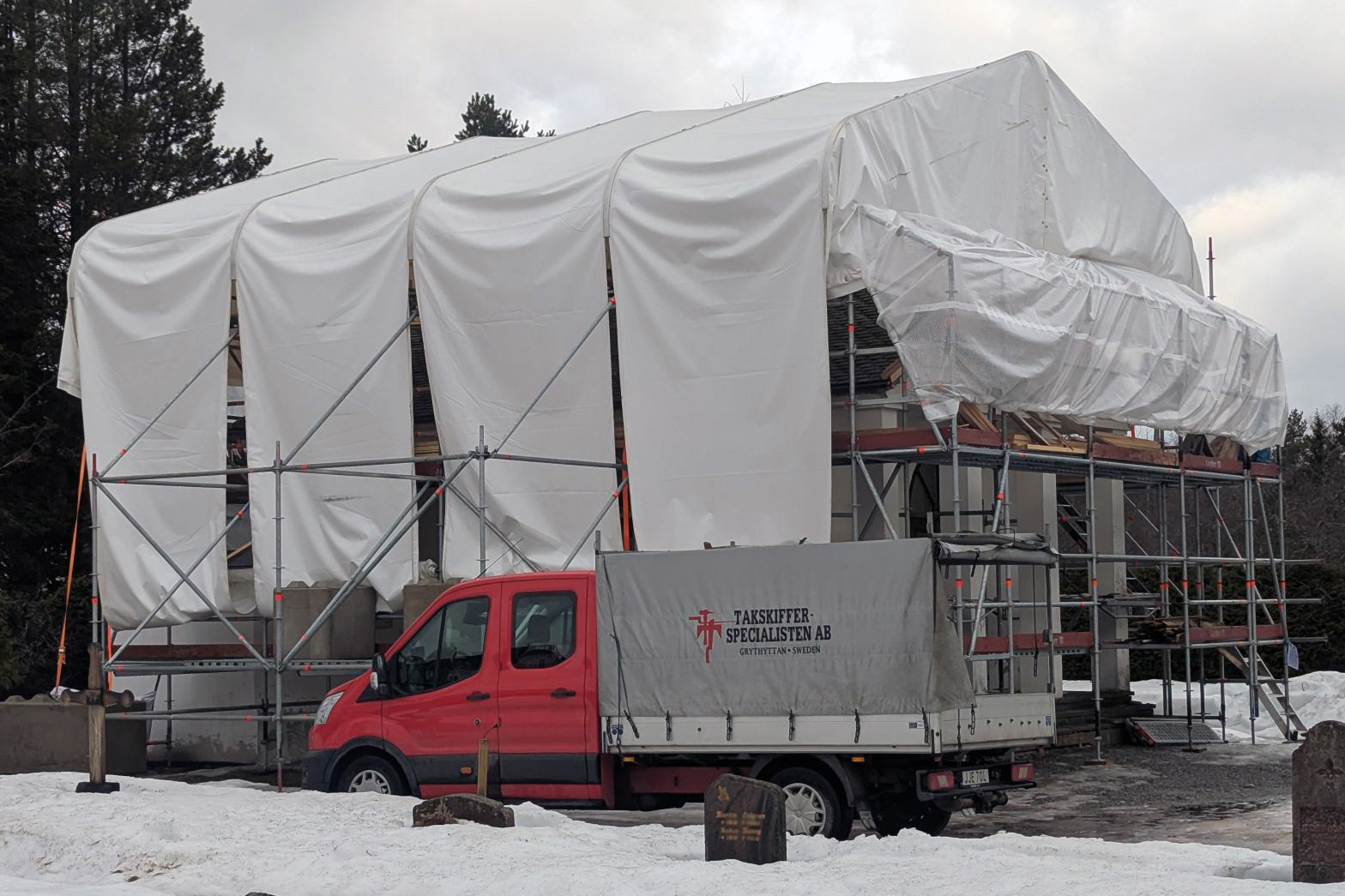 En byggnad täckt med byggställning och vit presenning. Byggnaden står på en kyrkogård, man skymtar några gravstenar Framför byggnaden står en pick-up med texten "takskifferspecialisten".