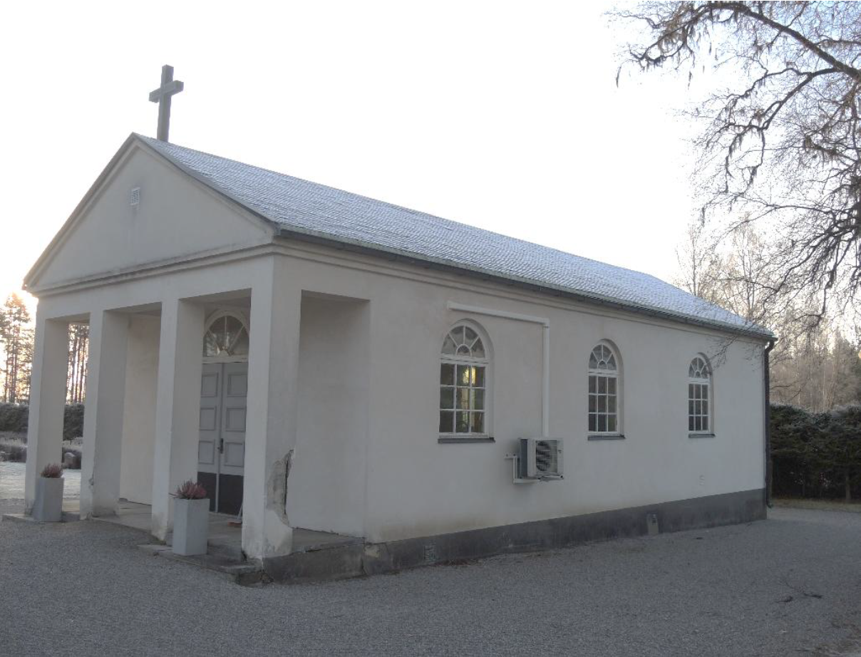 En raktangulär byggnad med ljus slätputsad fasad och skiffertak. På nocken ovan entrén sitter ett kors i koppar.