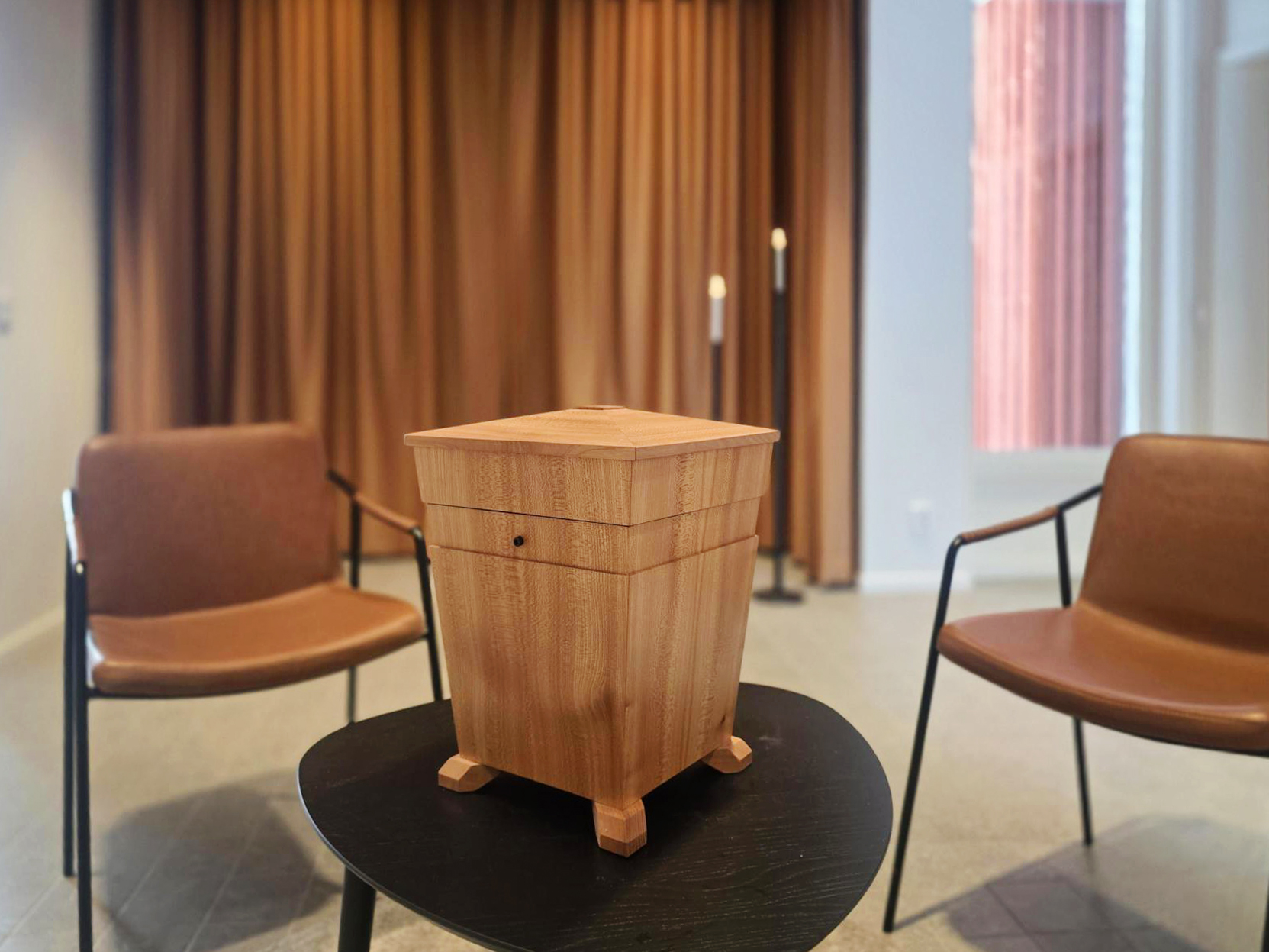En urna i trä som står på ett bort och med två stolar i bakgrunden.