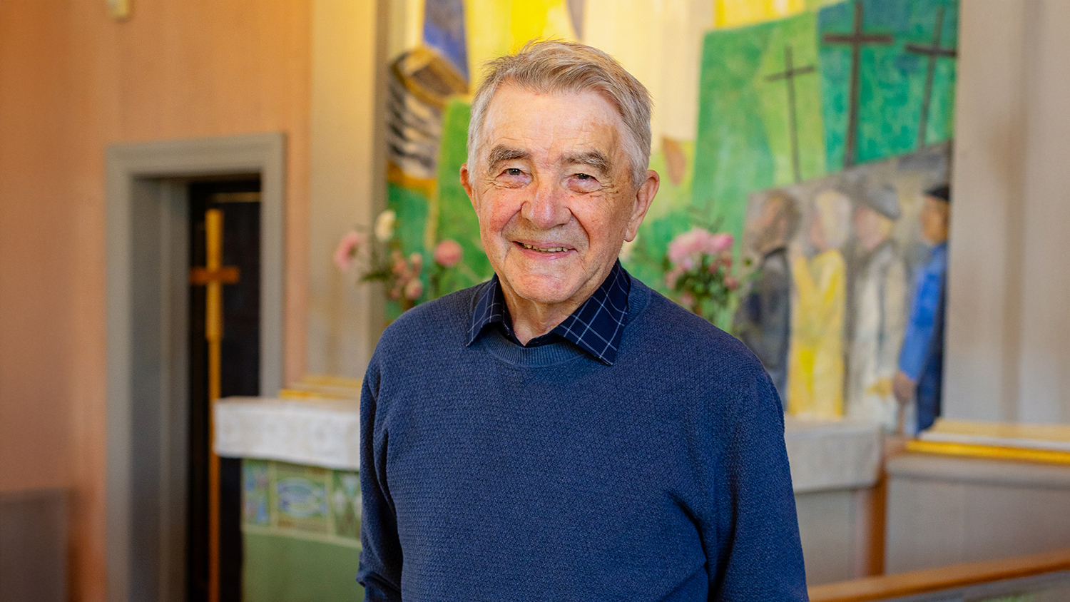 En äldre man som står i ett kyrkorum, med en altartavla i bakgrunden.