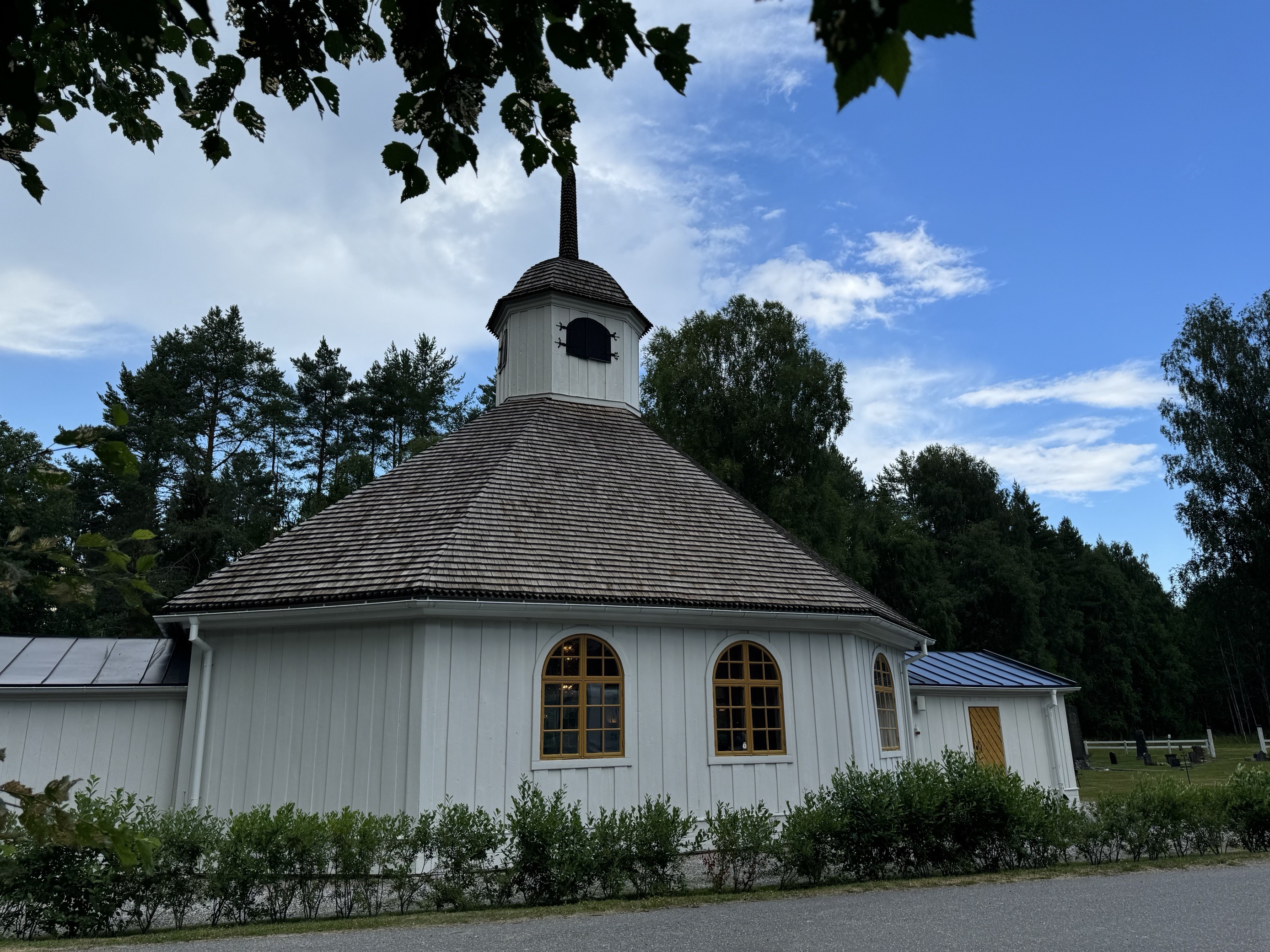 Vit träkyrka med brunt spåntak och en liten klockstapel med spetsigt torn. Kyrkan har rundbågiga fönster med gulmålade karmar och en gul dörr. Den ligger intill en väg, omgiven av grönska och höga träd, med en kyrkogård synlig i bakgrunden under en delvis molnig himmel.