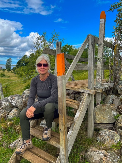 En kvinna sitter på en tre trappa som går över en sten gärsgård på ett fjäll. I förgrunden ses en stolpe med en orange markering. Himlen är blå och ett vitt fluffigt moln.