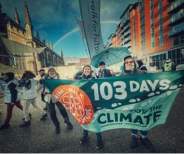 Klimatvandrare håller en banderoll med texten "103 DAYS FOR THE CLIMATE" på ett stadstorg framför historiska och moderna byggnader.