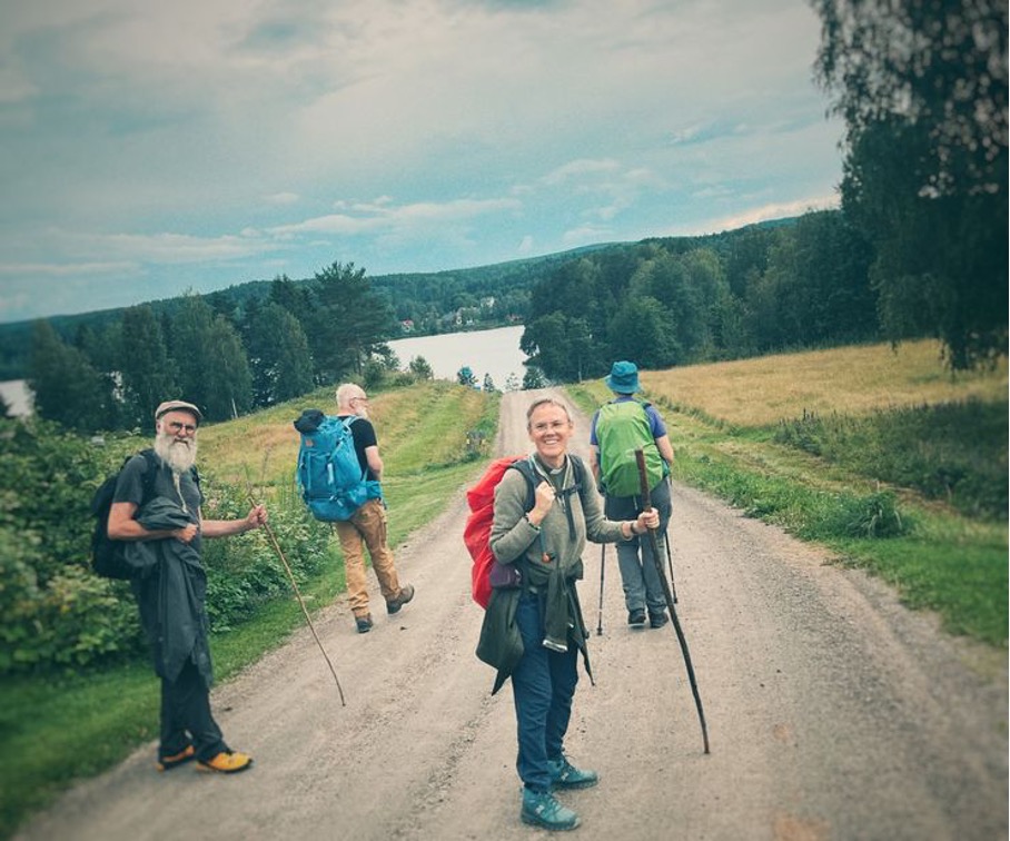 Kort alt-text (för skärmläsare) Fyra vandrare med ryggsäckar och vandringsstavar går längs en grusväg på landsbygden, med en sjö och gröna skogar i bakgrunden.