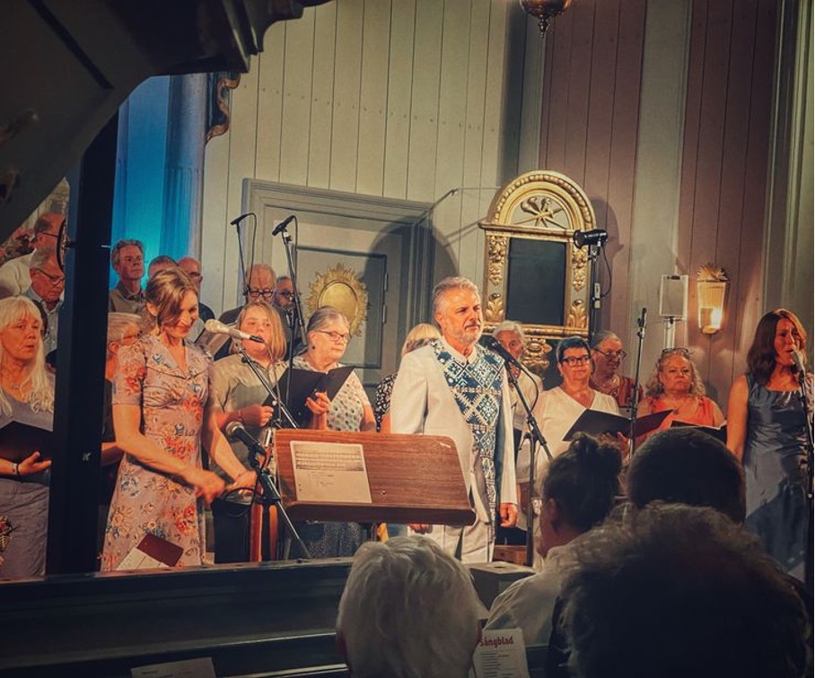 En kör och solister uppträder vid mikrofoner inne i en kyrka framför ett trästativ med noter.