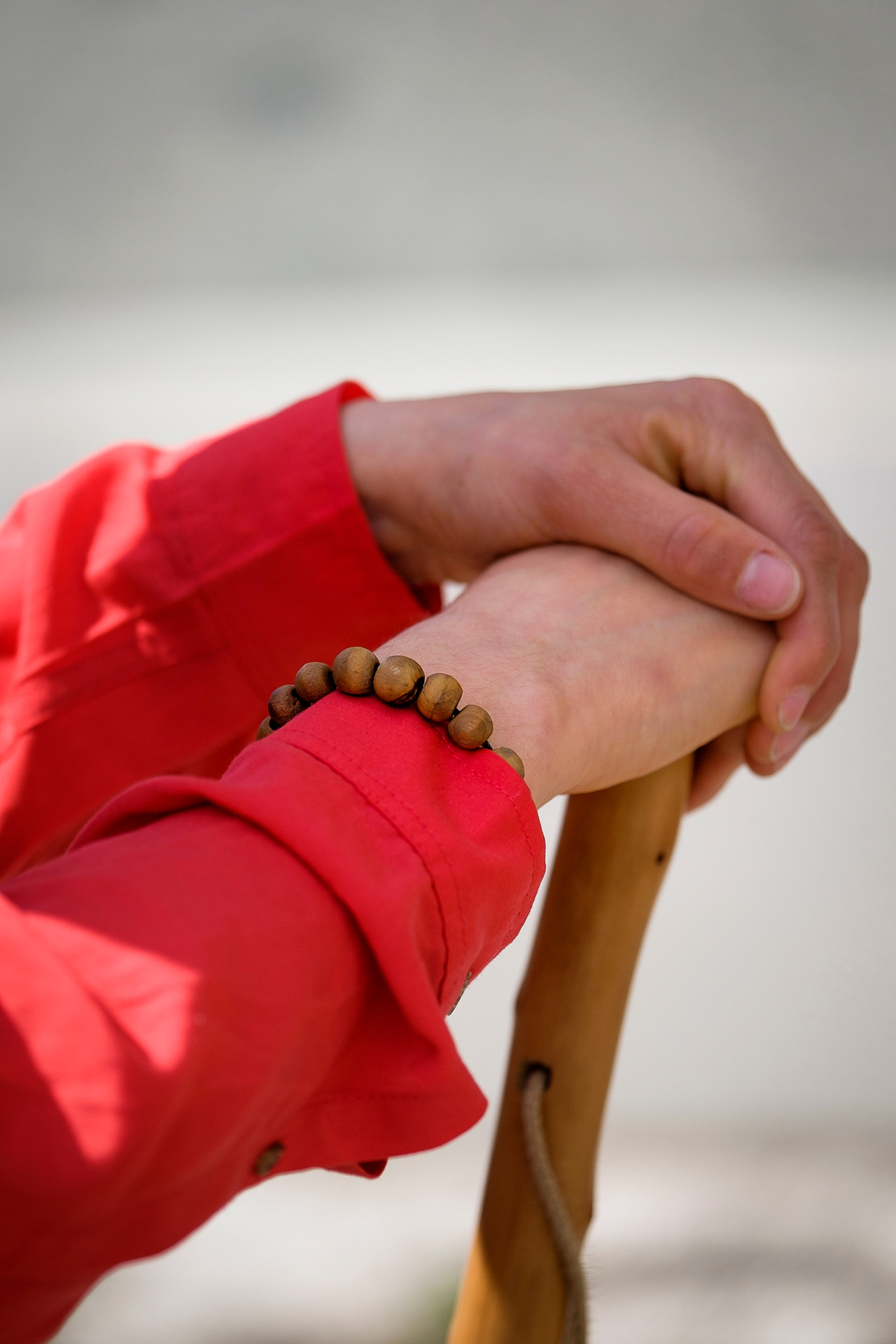 Händer som vilar mot en käpp. Röd skjorta och radband runt handleden.