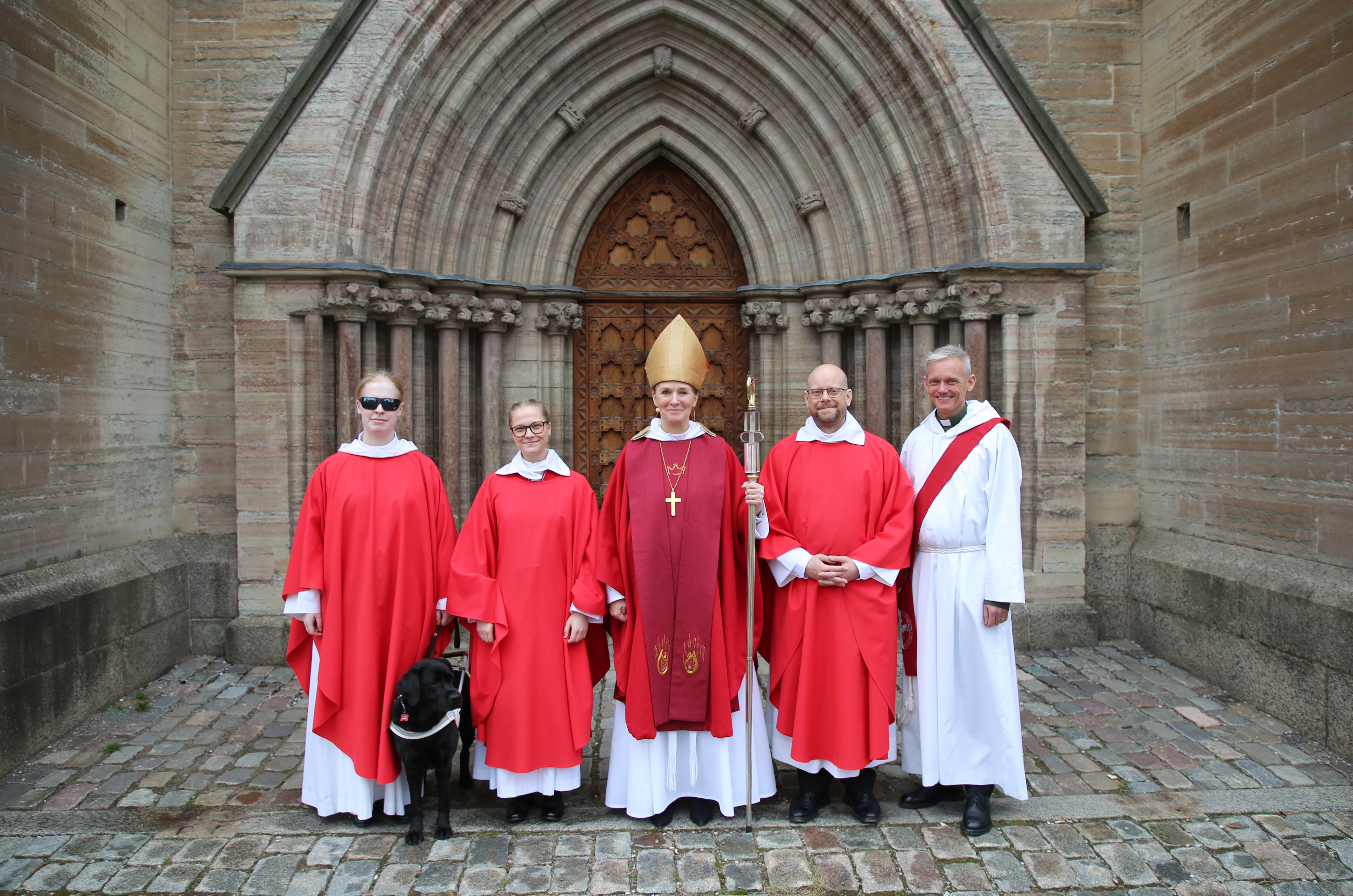 Tre nyvigda präster, en nyvigd diakon, en ledarhund och en biskop står utomhus framför en kyrkport.