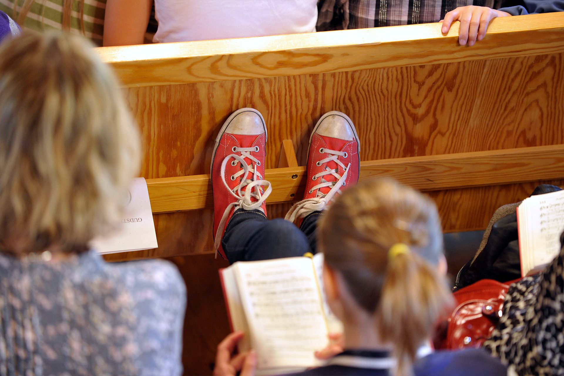 En kyrkbänk sedd bakifrån. Ett barn tittar i en psalmbok och har fötterna på bänken framför.