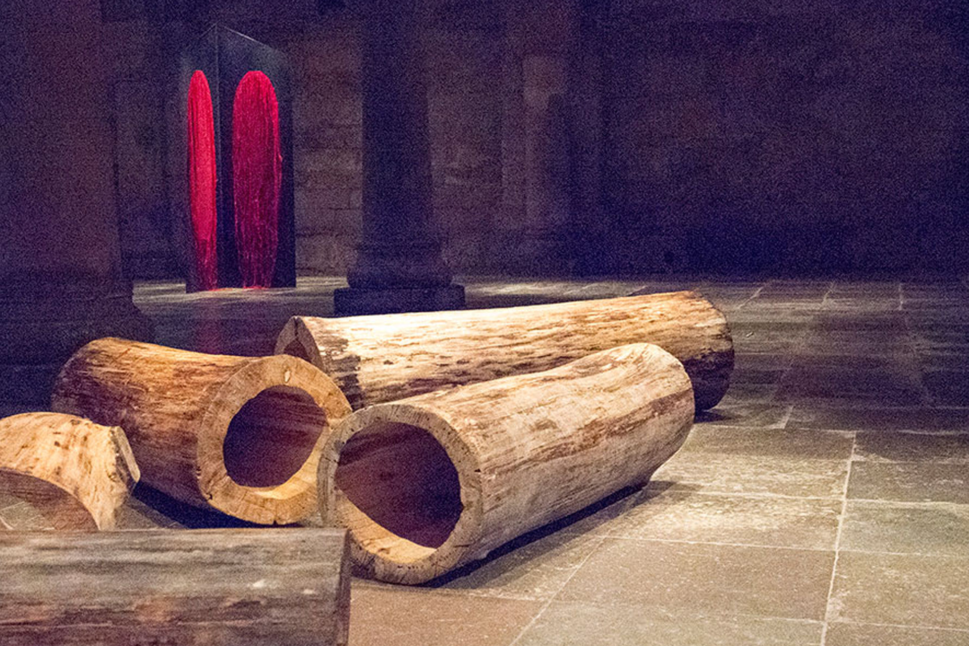 I förgrunden syns urholkade, liggande trädstammar. I bakgrunden skymtas en mörk konstruktion med två valv täckta av röda draperier.