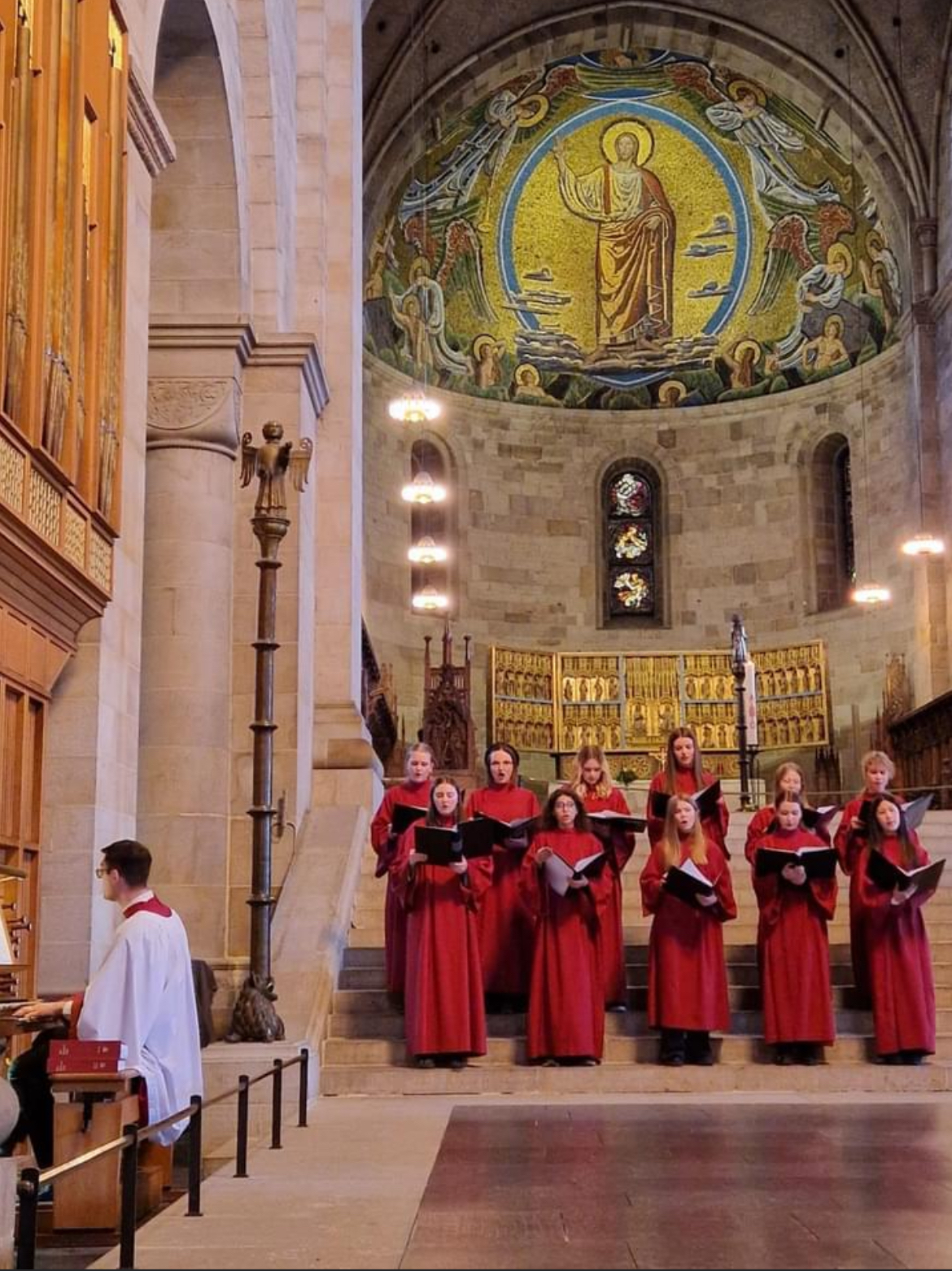 En grupp flickor klädda i röda kåpor med pärmar i händerna sjunger i Domkyrkan. Till vänster sitter en vitklädd person framför en orgel och ackompanjerar.