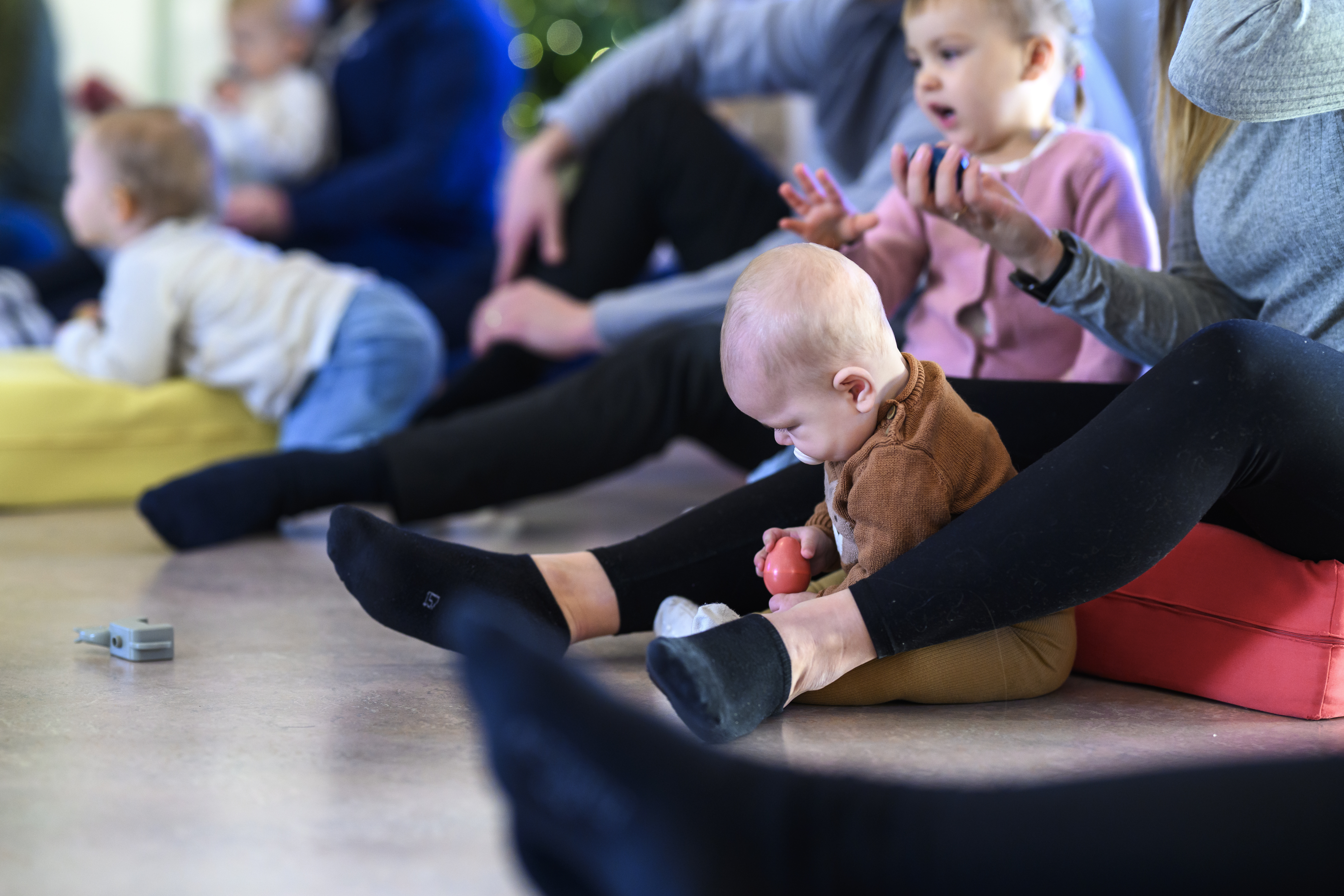 En baby sitter på golvet och leker meden boll. Runt omkring ser man syskon och föräldrar som sjunger.