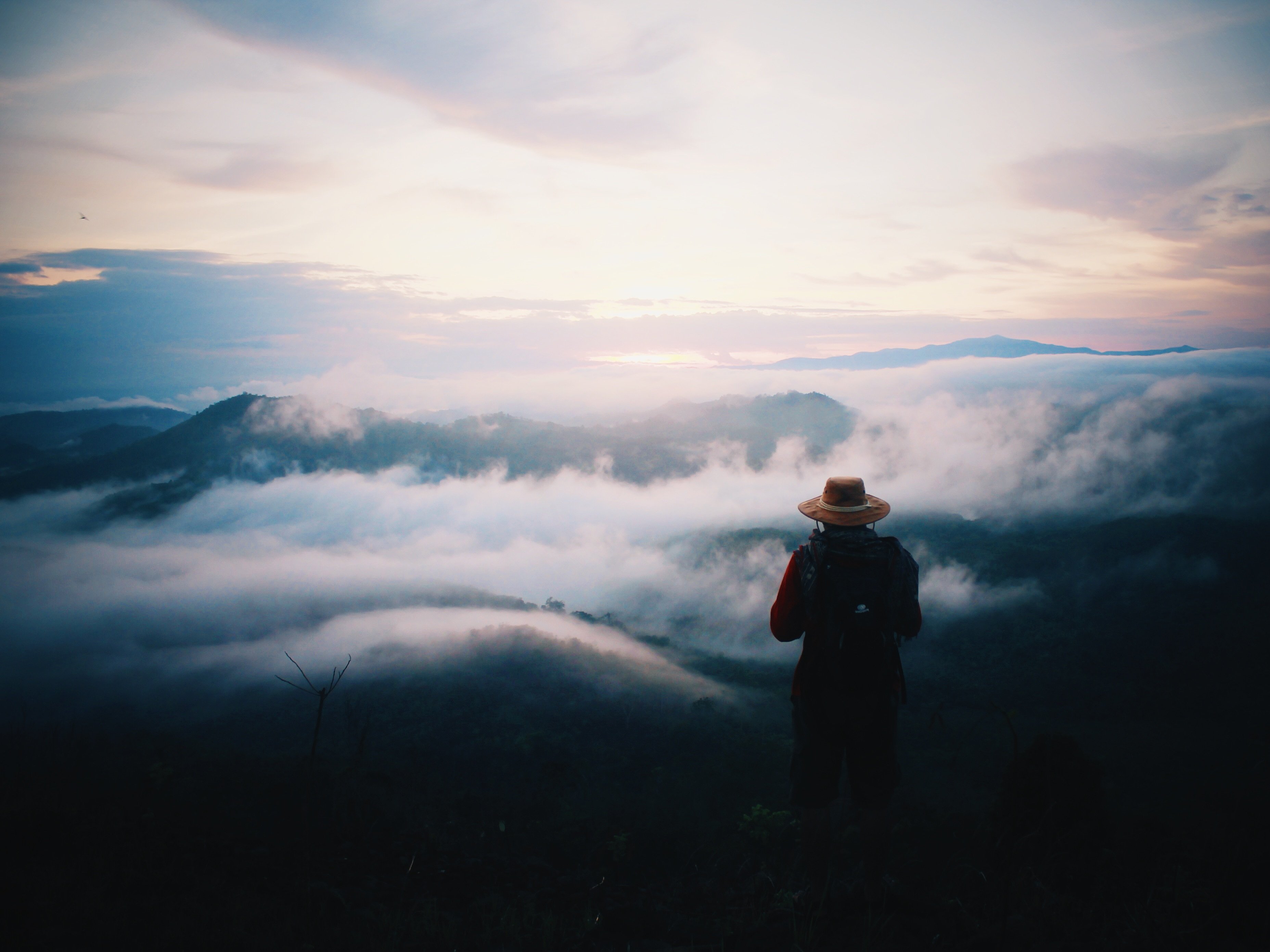 Person mit Hut blickt von Berggipfel herunter auf die Wolken im Tal