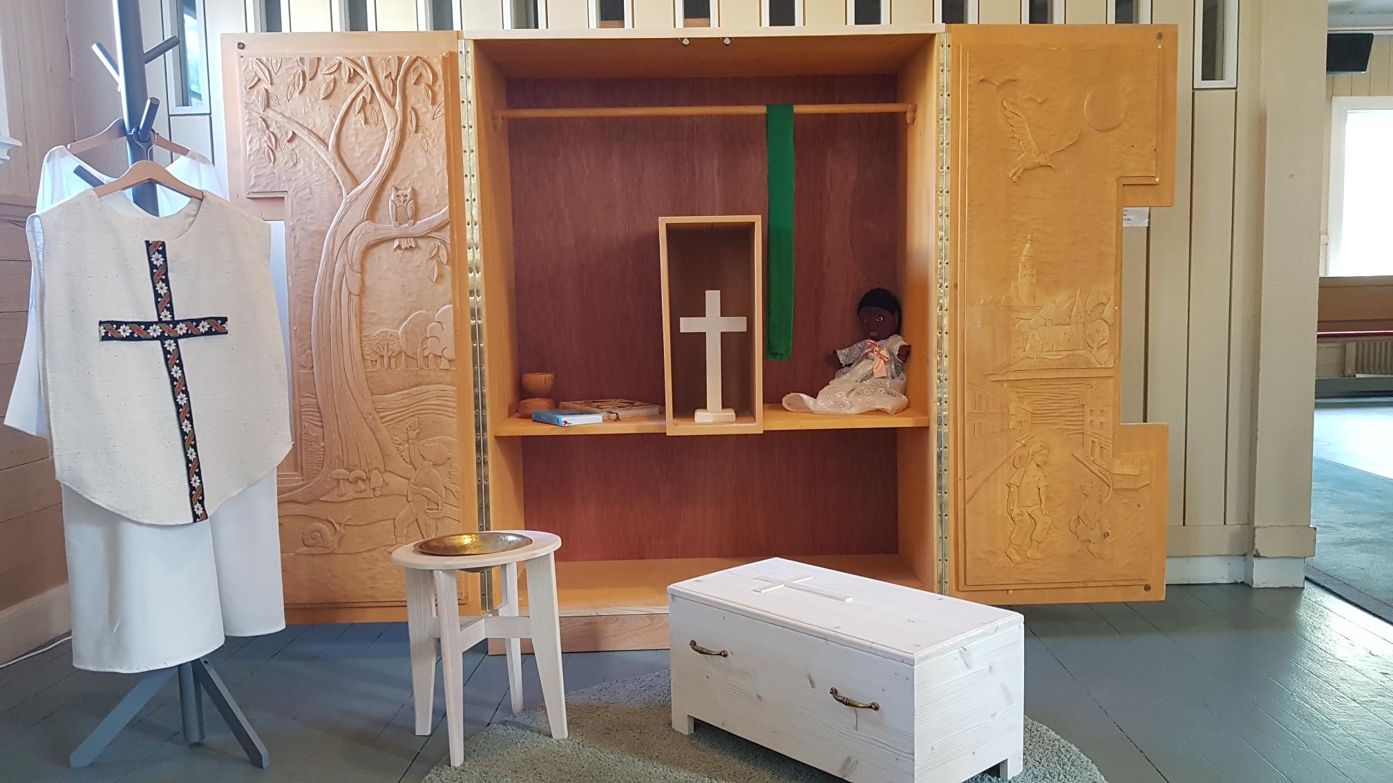Ett altarskåp i barnstorlek med dörrarna öppna. Insidan av dörrarna är täckta med bildhuggerier. I skåpet finns ett kors, en docka, några böcker och två träkärl för nattvardslek. Utanför skåpet står en liten vit kista, en liten dopfunt och en klädhängare med prästkläder. 