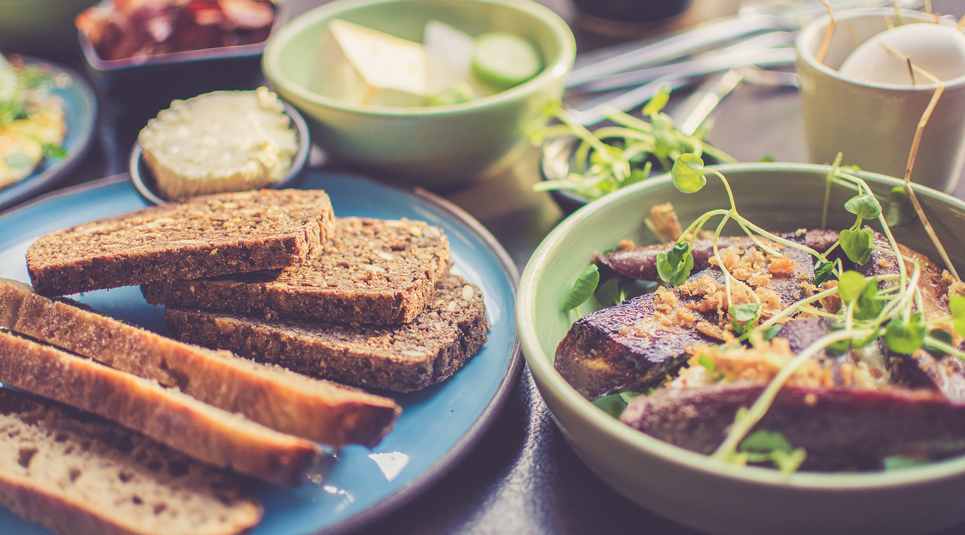 Tallrikar och skålar med grov skivat bröd i förgrunden. I bakgrunden skålar med ägg och grönsaker.