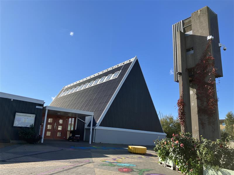 En grå träbyggnad, före detta kyrka, där den före detta kyrksalen är byggd som en stor trekant. I förgrunden står en klockstapel i betong och några odlingslådor med blommor.  