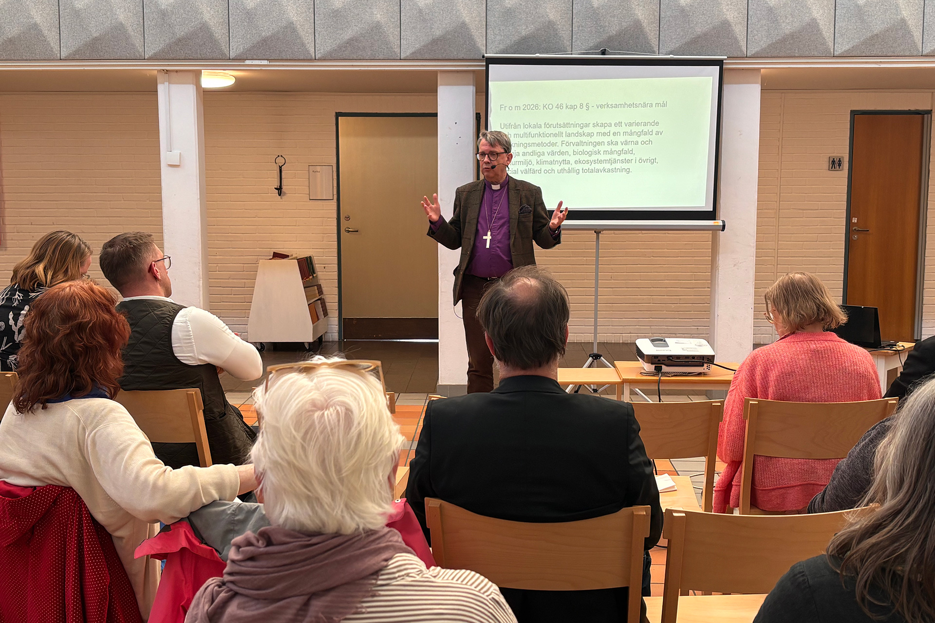 En man i lila prästskjorta står längre fram i en samlingssal och talar till en sittande publik. Bakom honom finns en projektorduk med text, och i rummet syns stolar, en projektor på bord och ljusa tegelväggar.