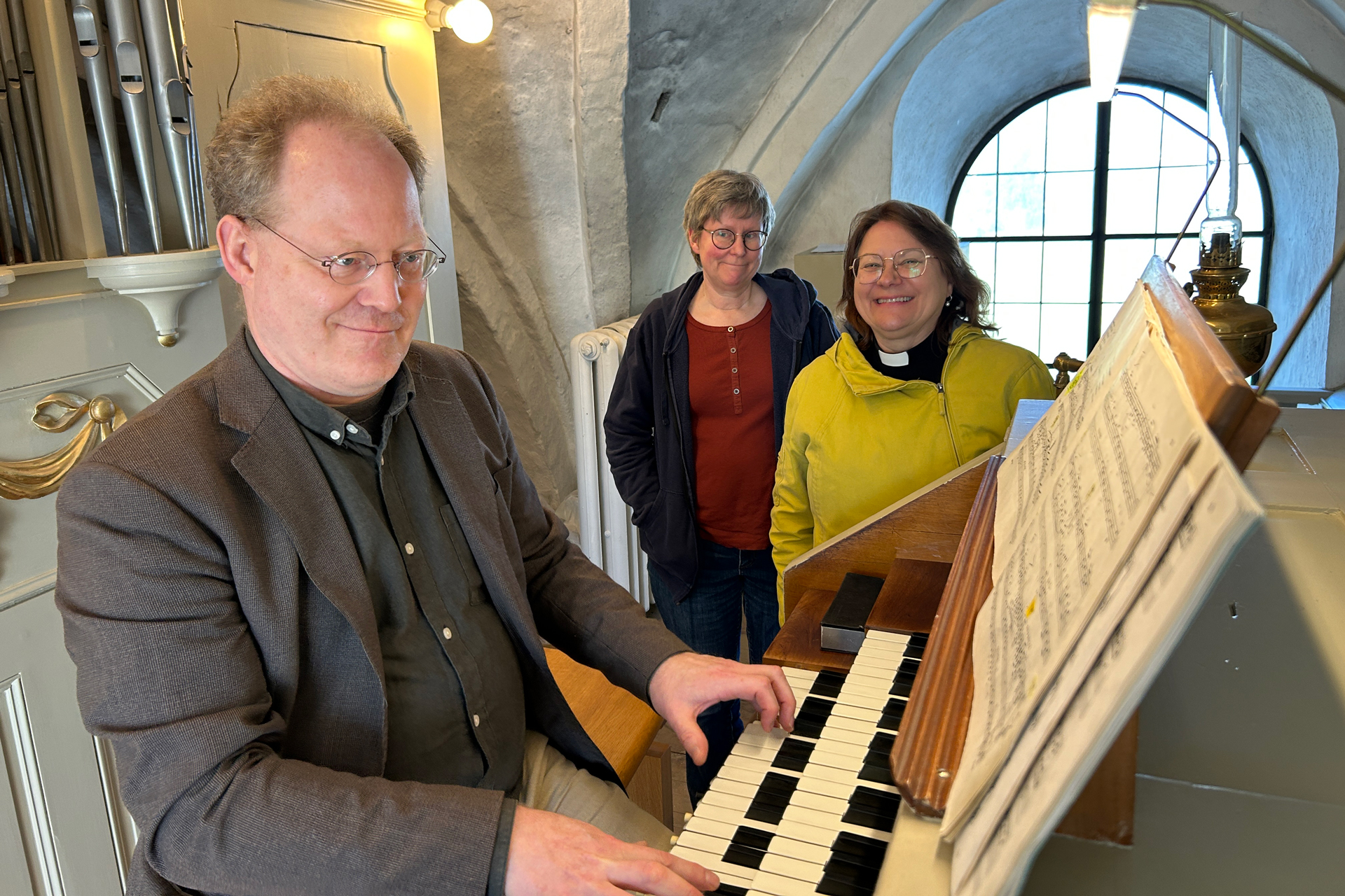 En man spelar på orgeln på orgelläktaren. Bredvid honom står två leende kvinnor.
