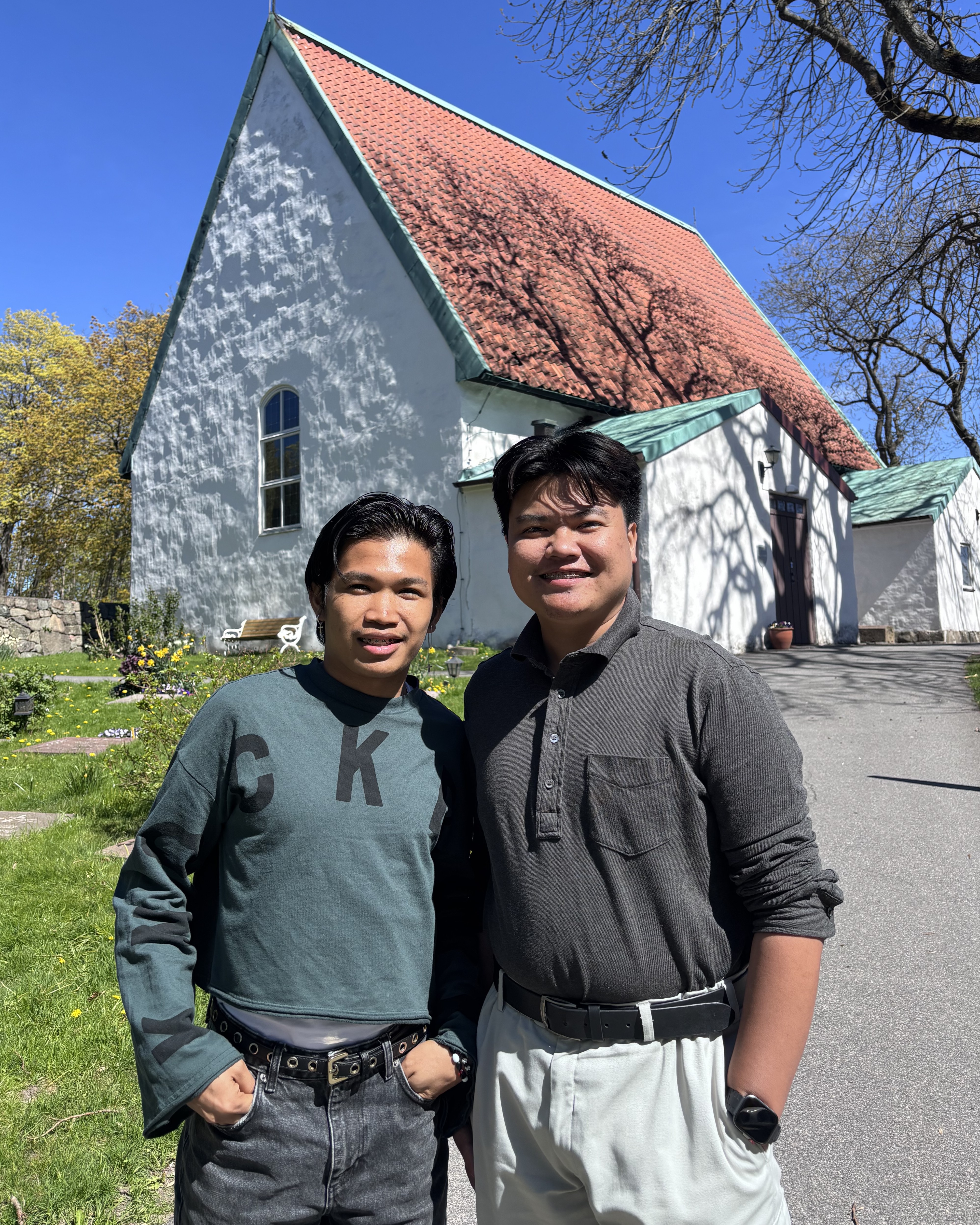 Två unga män ler mot kameran framför en solig Lundby gamla kyrka.
