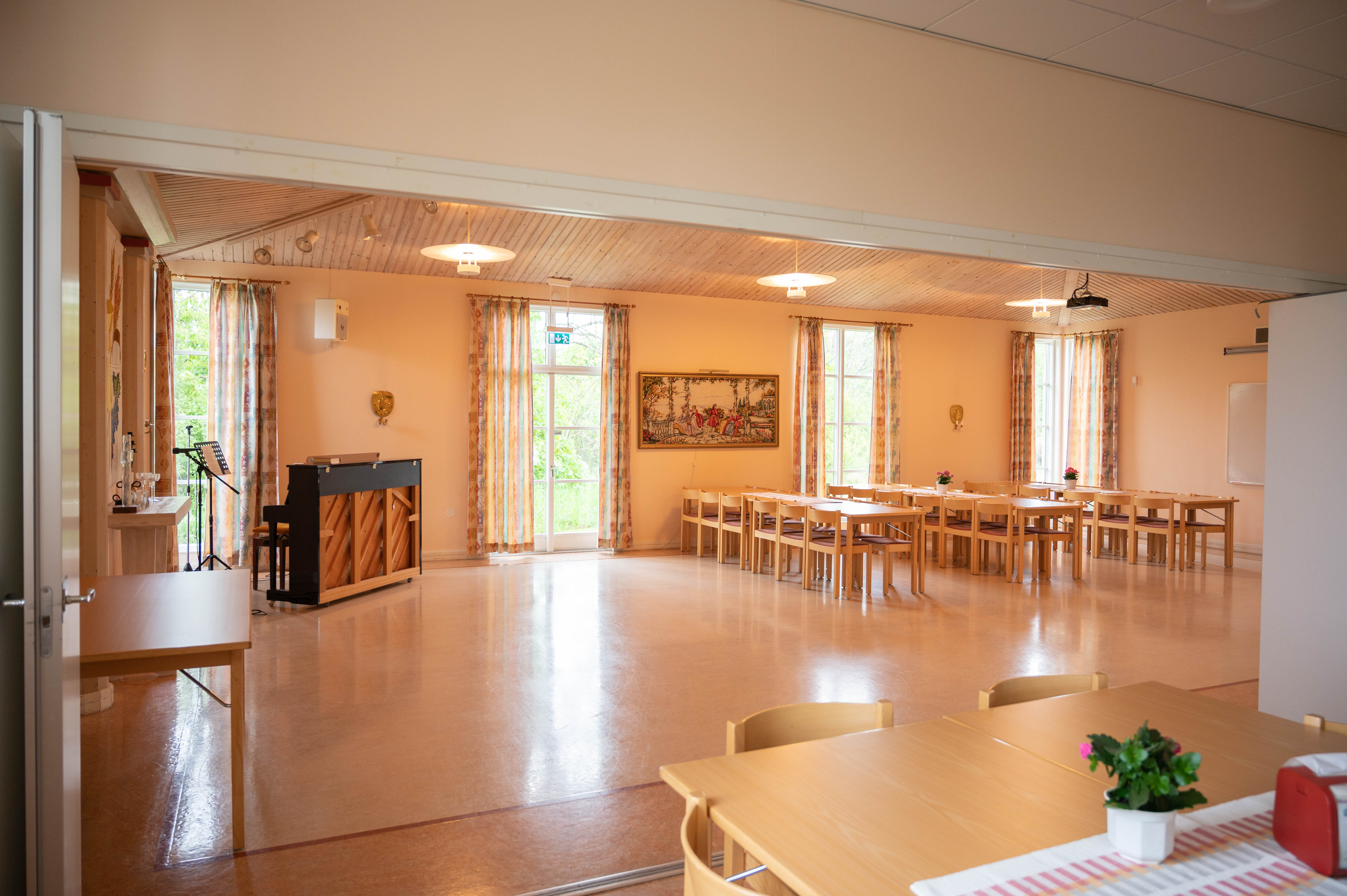 Vy över samlingssalen i Kimstads församlingshem. Till vänster står ett piano och till höger står långbord med stolar uppställda.