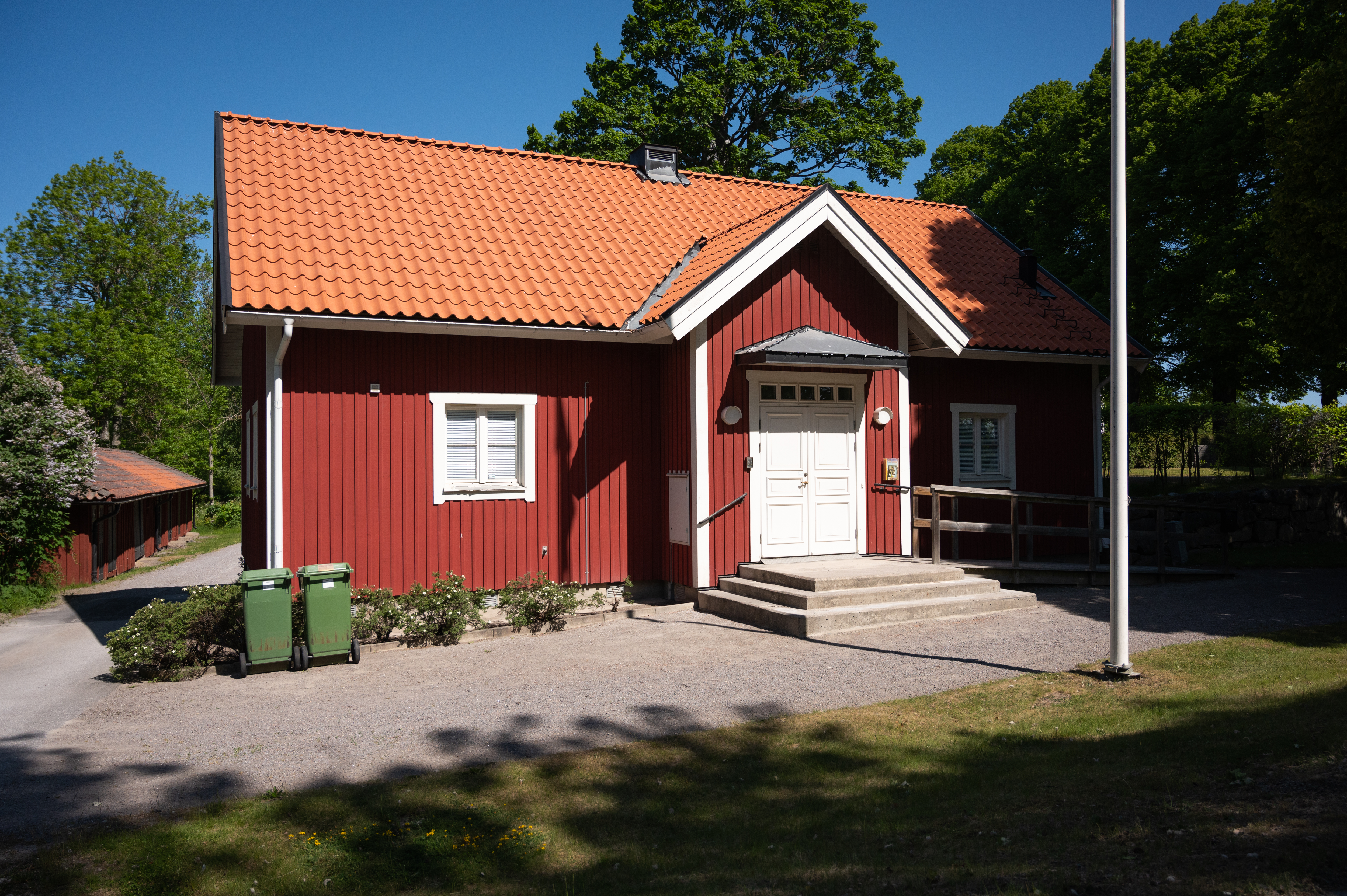 En röd stuga med tegeltak syns i mitten av bilden. Till höger syns en flaggstång.  Runt stugan syns grönska och i bakgrunden syns ett kyrktorn.