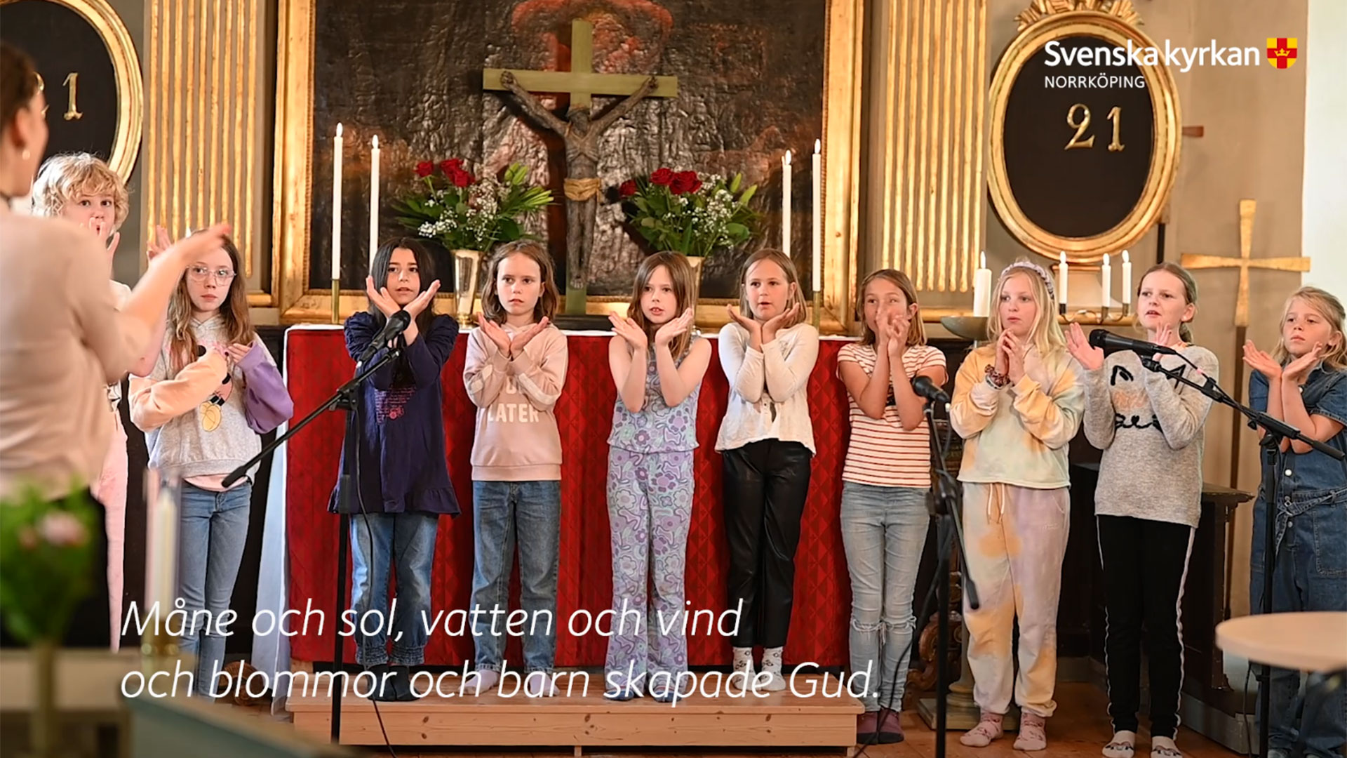 Vy över ett kor i kyrkan där en barnkör står uppställd på rad och sjunger och gör rörelser till musiken.