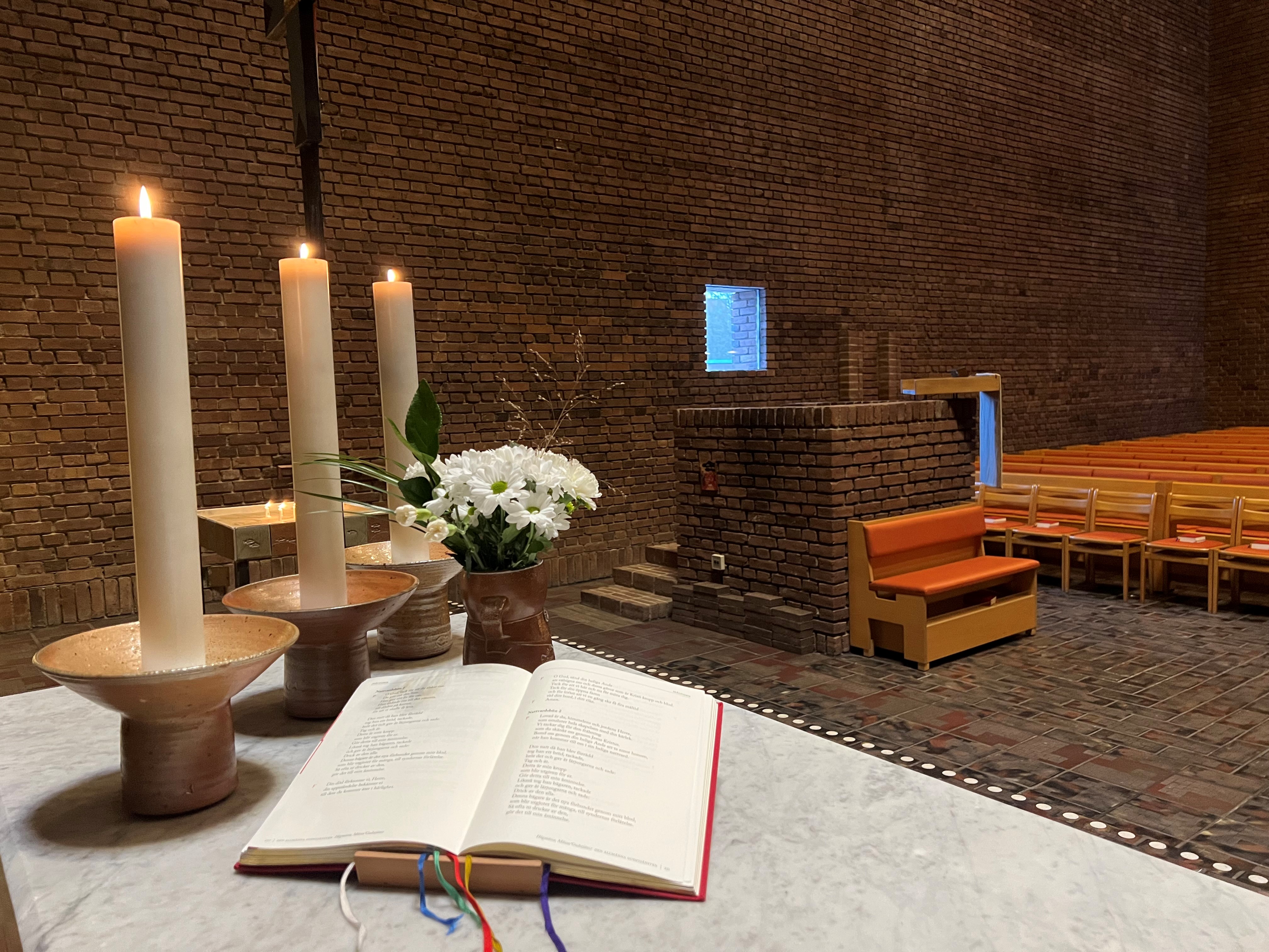 Del av altare i kyrkan med ljus, blommor och bok. Ljusbärare och predikstol i bakgrunden.