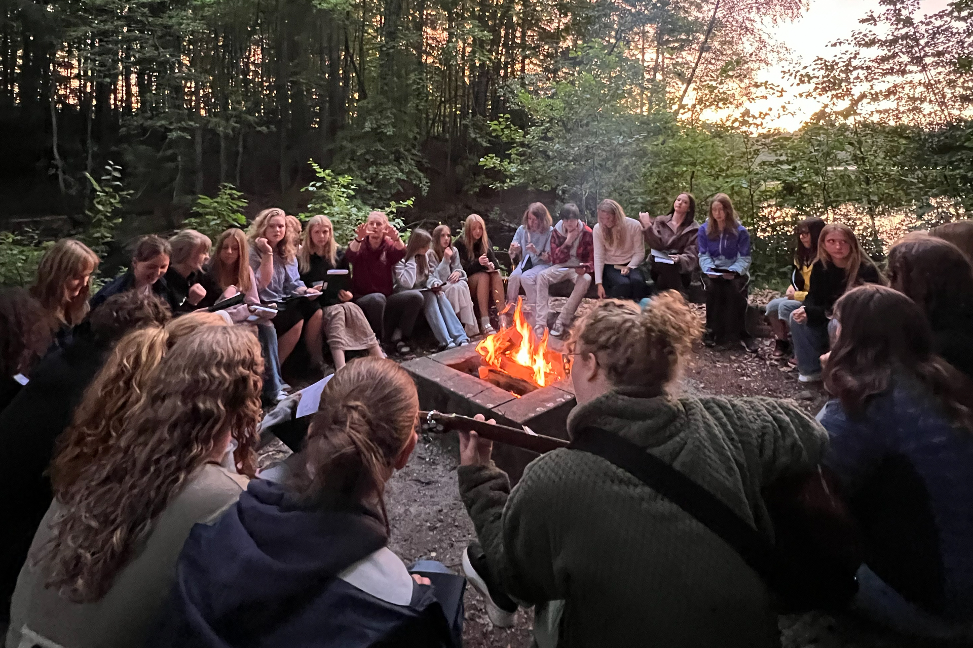 En stor grupp ungdomar sitter runt en lägereld. En person spelar gitarr. 