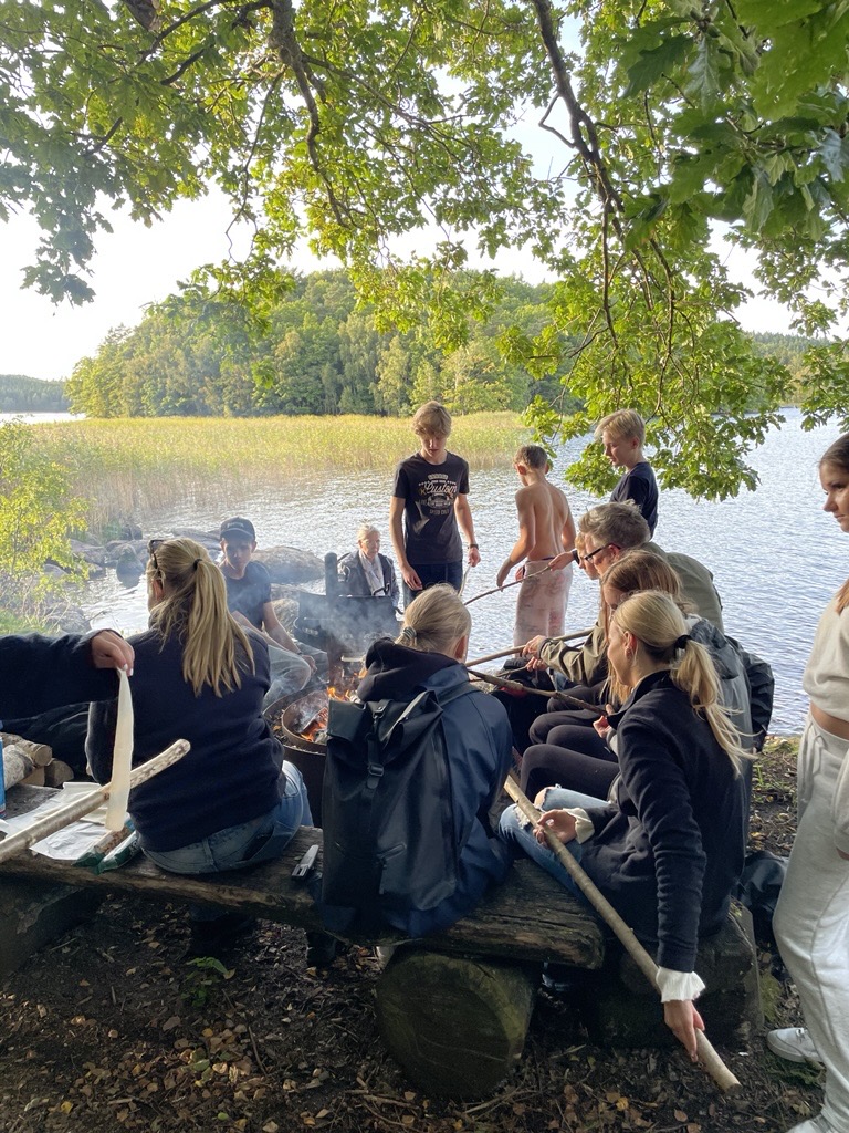 Ungdomar står och sitter runt en lägereld. Det är vid en sjö och det ser ut att vara på sommaren. 