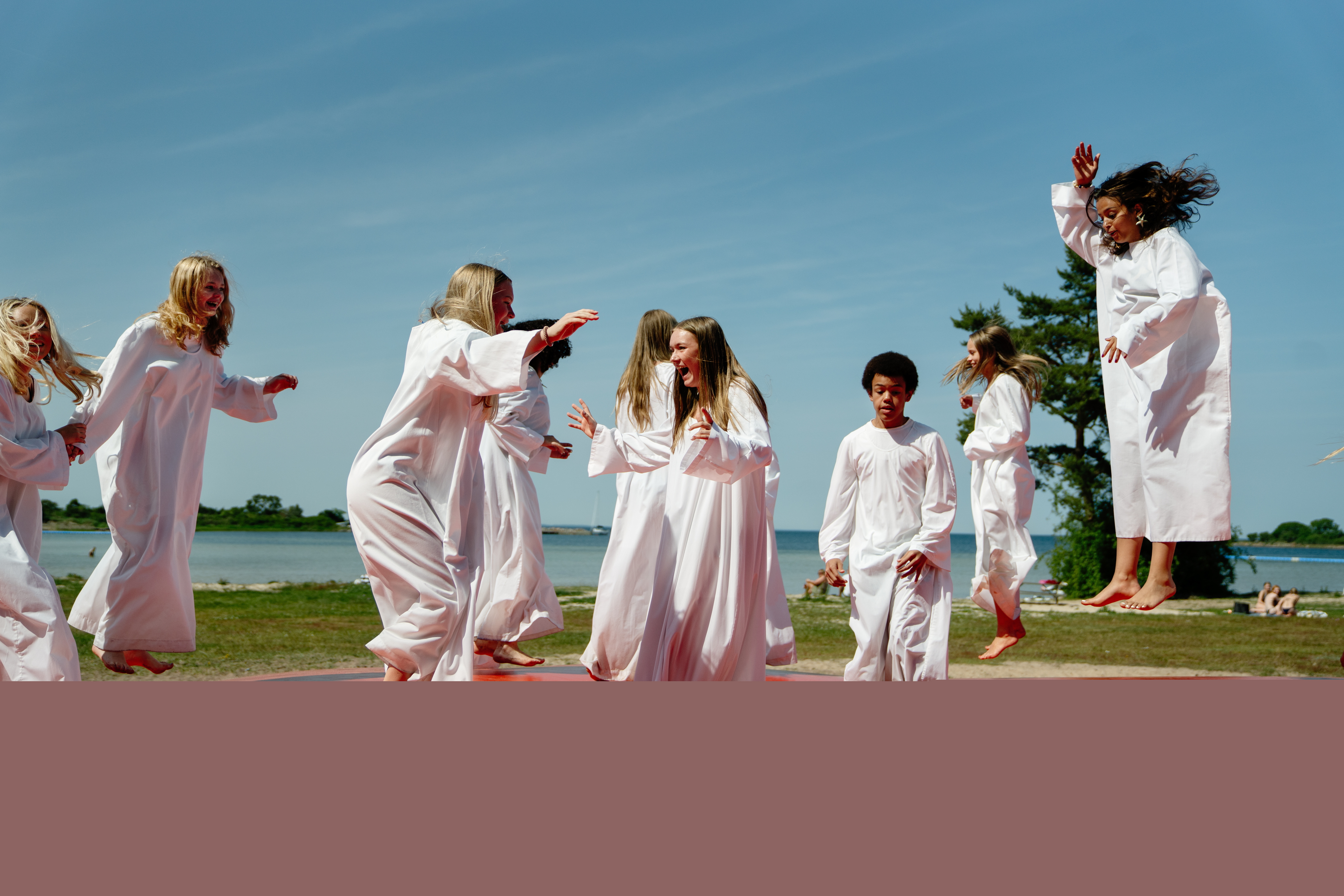 Åtta konfirmander klädda i vita kåpor hoppar på en studsig madrass på en strand under sommartid. De har väldigt roligt.