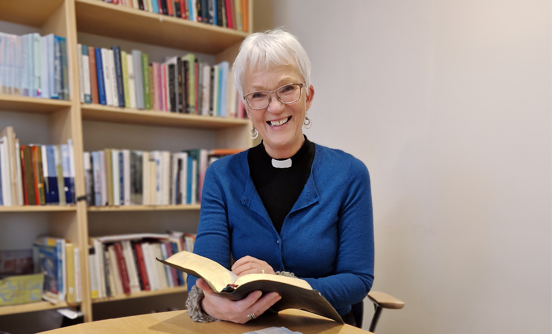 Leende kvinna med kort vitt hår, blå kofta och prästkrage runt halsen i svart skjorta sitter framför suddig bokhylla och håller i en bok