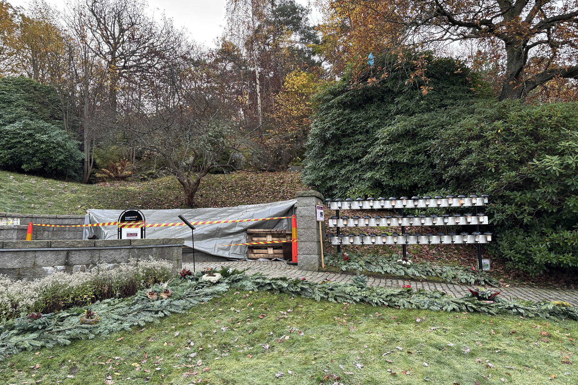 Minneslund med stenmurar, stenlagd gång, ljusbärare och en presenning med gulröda avspärningsband framför. I bkagrunden buskar, träd och gräs i höstfärger.