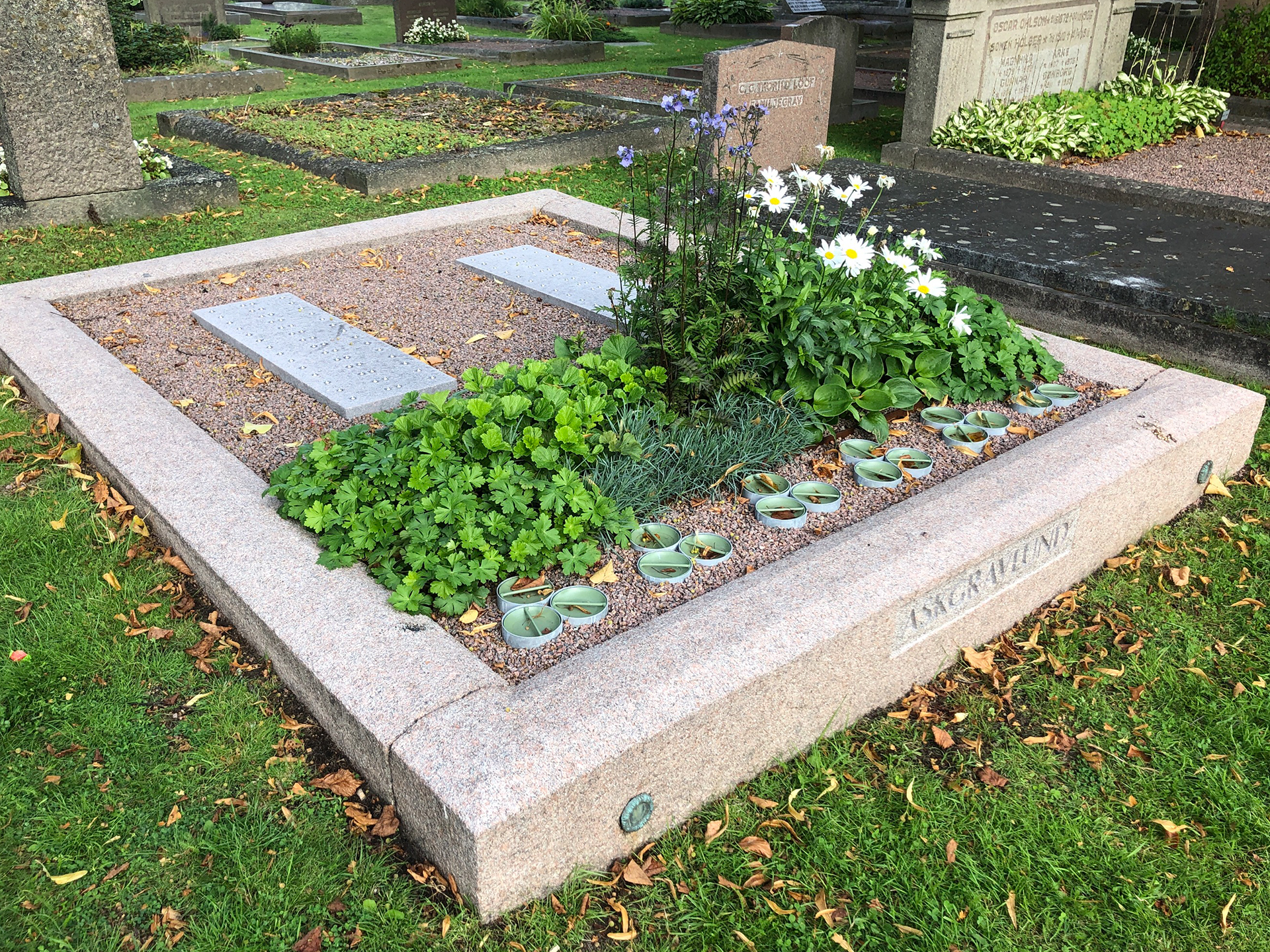 Gravplats med stenram och planteringsyta med växter. Belagd med grus vari två stenplattor ligger.