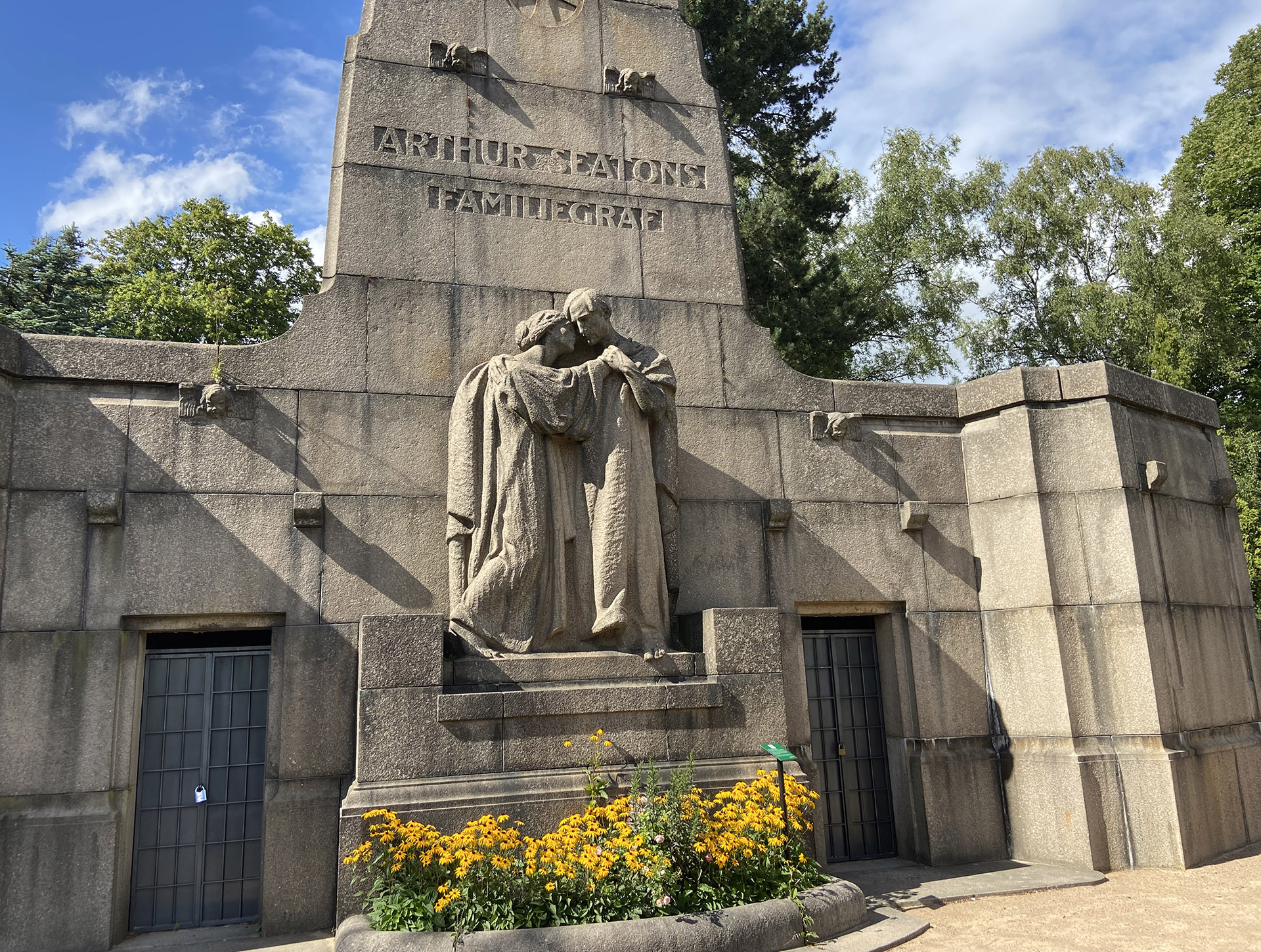 Stort gravmonument i sten med skulptur, inskription Arthur Seatons familiegraf med gula blommor framför