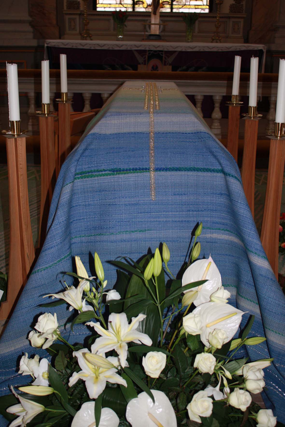 Bårtäcke över en kista med ljus omkring och vita liljor.